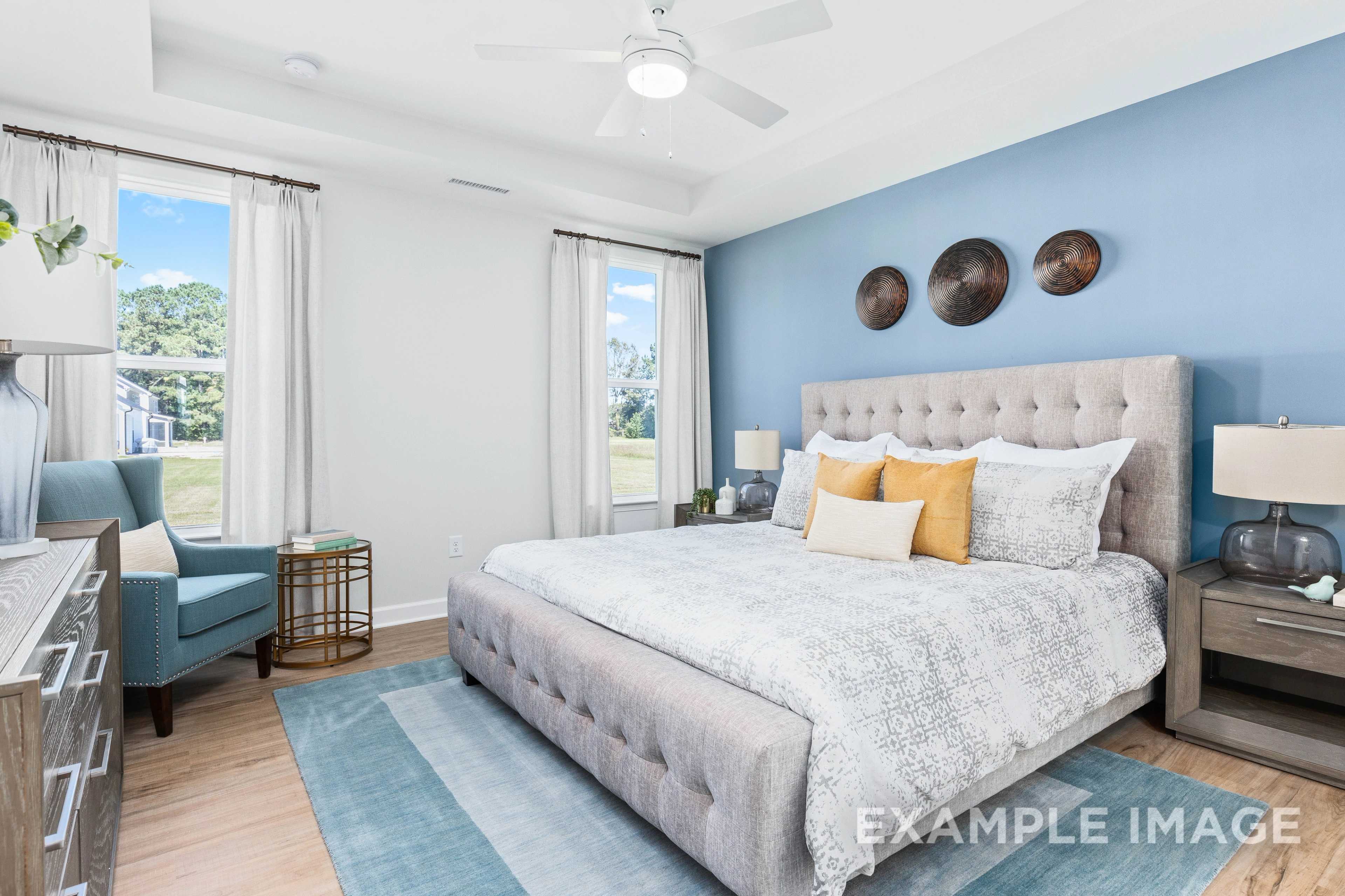Spacious master bedroom in The Ash C with blue accent wall, tufted king bed, yellow pillows, armchair, and large sunny windows