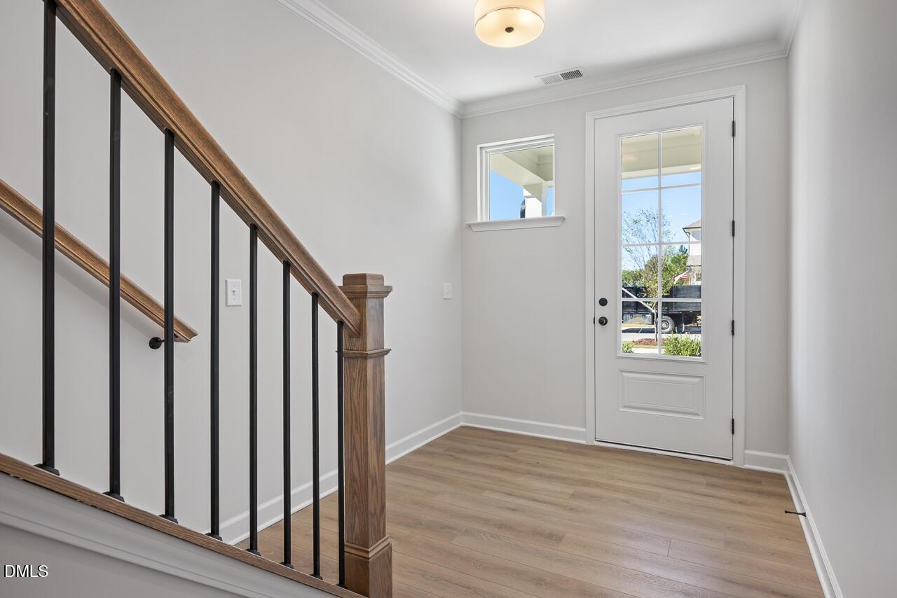 Bright foyer with glass-panel front door, wood-look flooring, and oak staircase with iron balusters in The Preston C, Wendell Ridge, NC