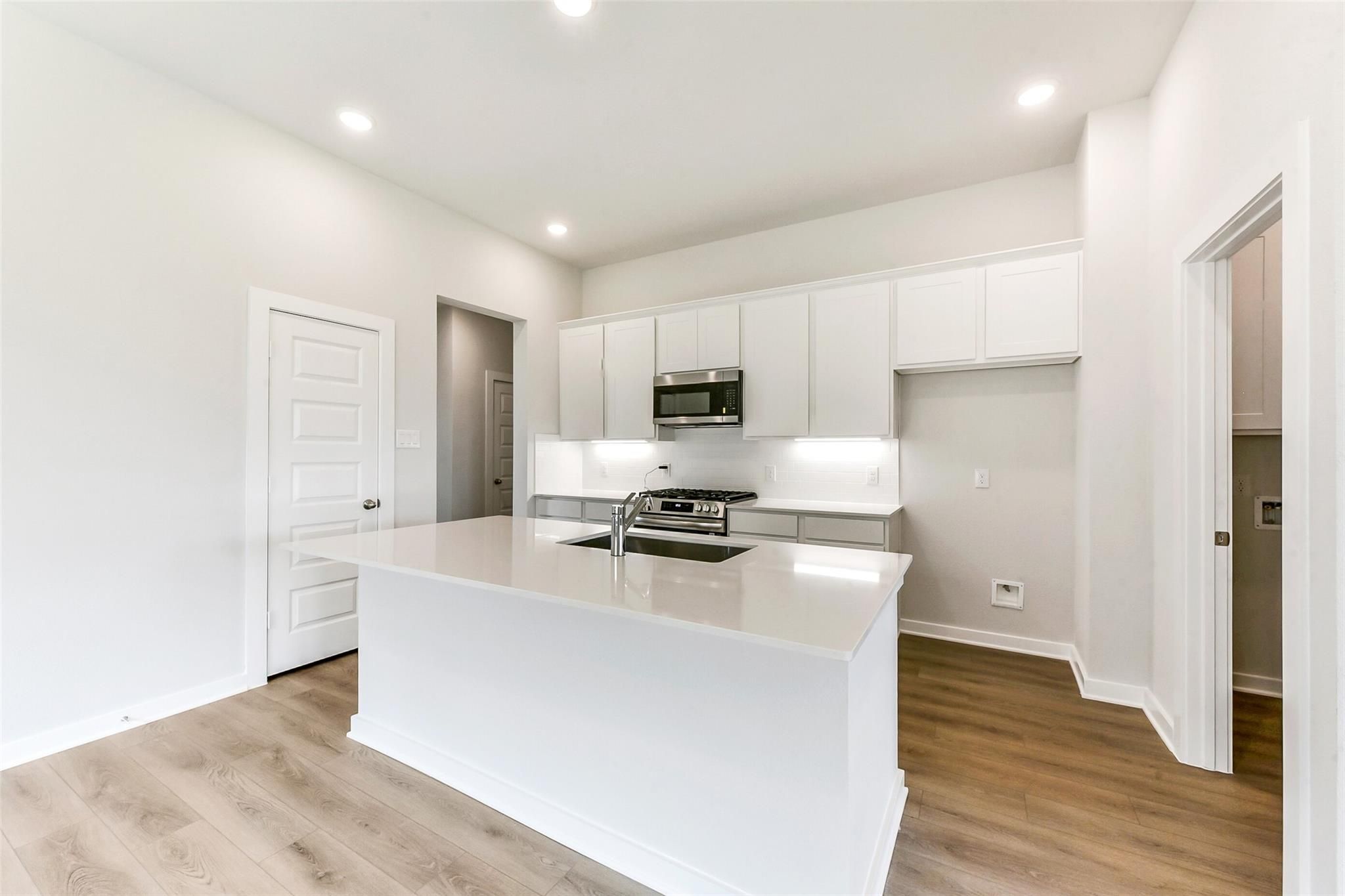 Modern white kitchen with large island, stainless appliances, and light wood floors in Davidson Homes The Costa B, Dayton, Texas