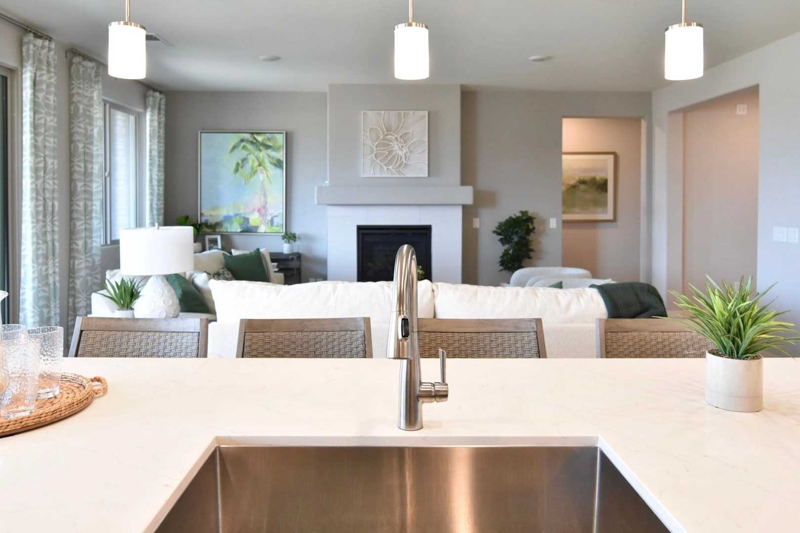 Open-concept kitchen in The Harmony Davidson Homes with white quartz island, stainless sink, woven stools, and adjacent living area fireplace
