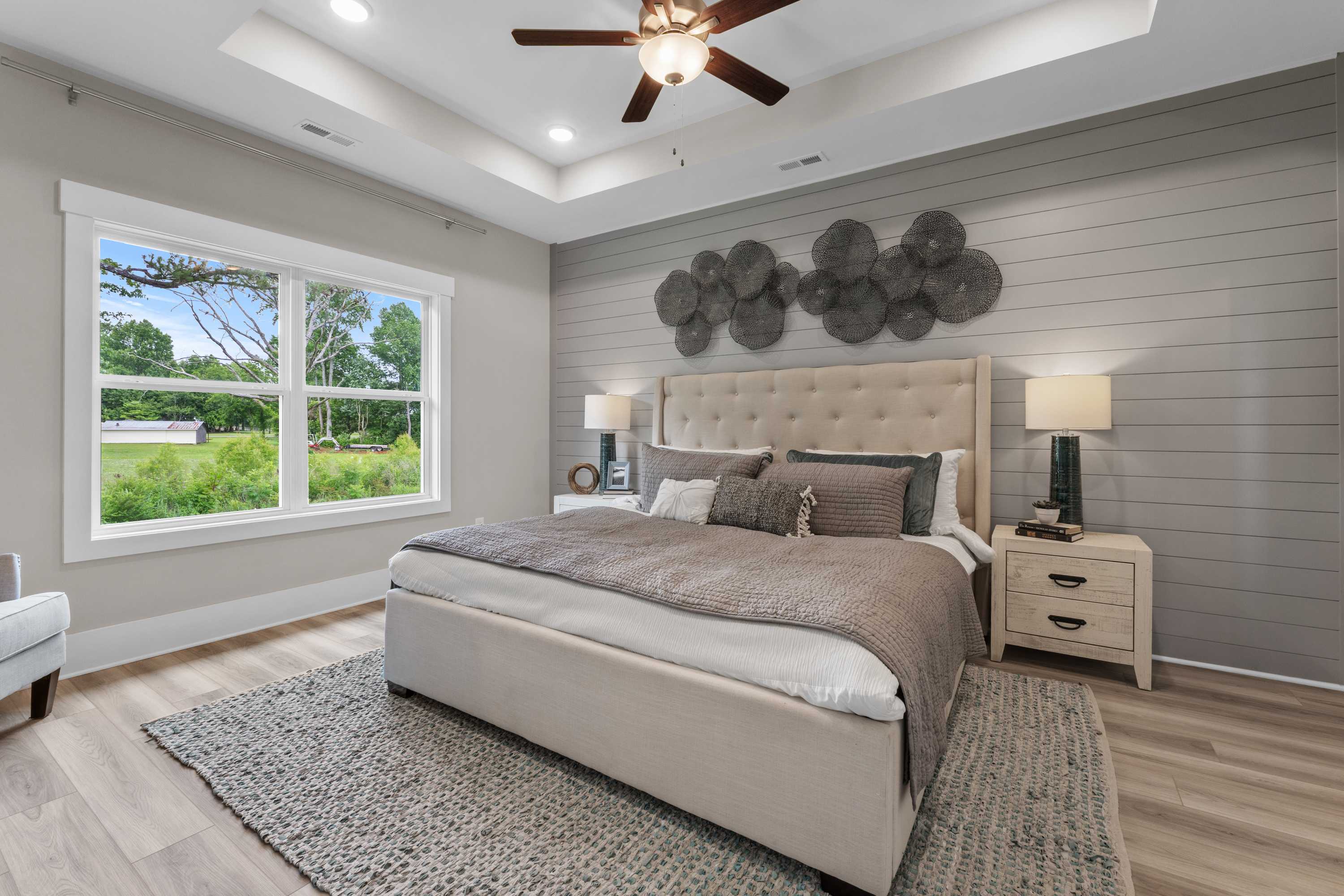 Spacious master bedroom at The Highlands in Arab Alabama by Davidson Homes with beige tufted bed, gray shiplap walls, ceiling fan, and lush window views