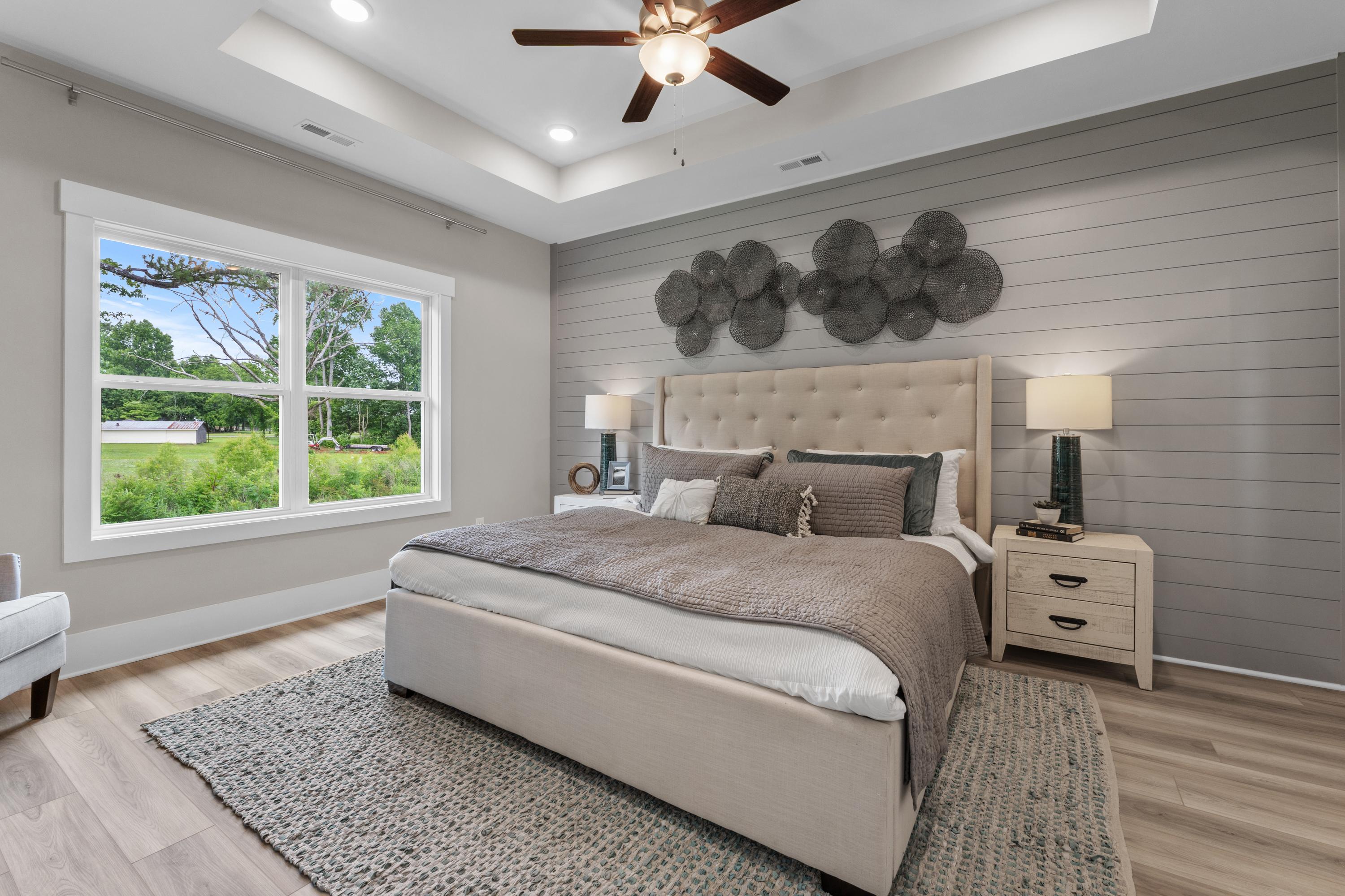 Spacious master bedroom at The Highlands in Arab Alabama by Davidson Homes with beige tufted bed, gray shiplap walls, ceiling fan, and lush window views