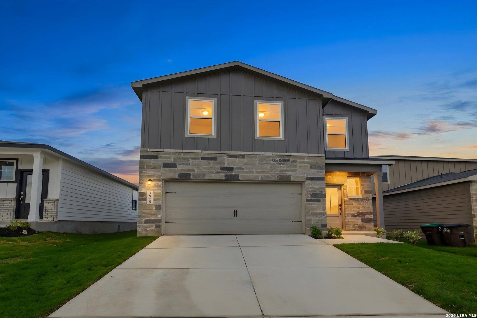 Modern 2-story gray home with stone facade, 2-car garage, lit windows, driveway, and green lawn at dusk in Agave, San Antonio TX - Davidson Homes The Blanco C