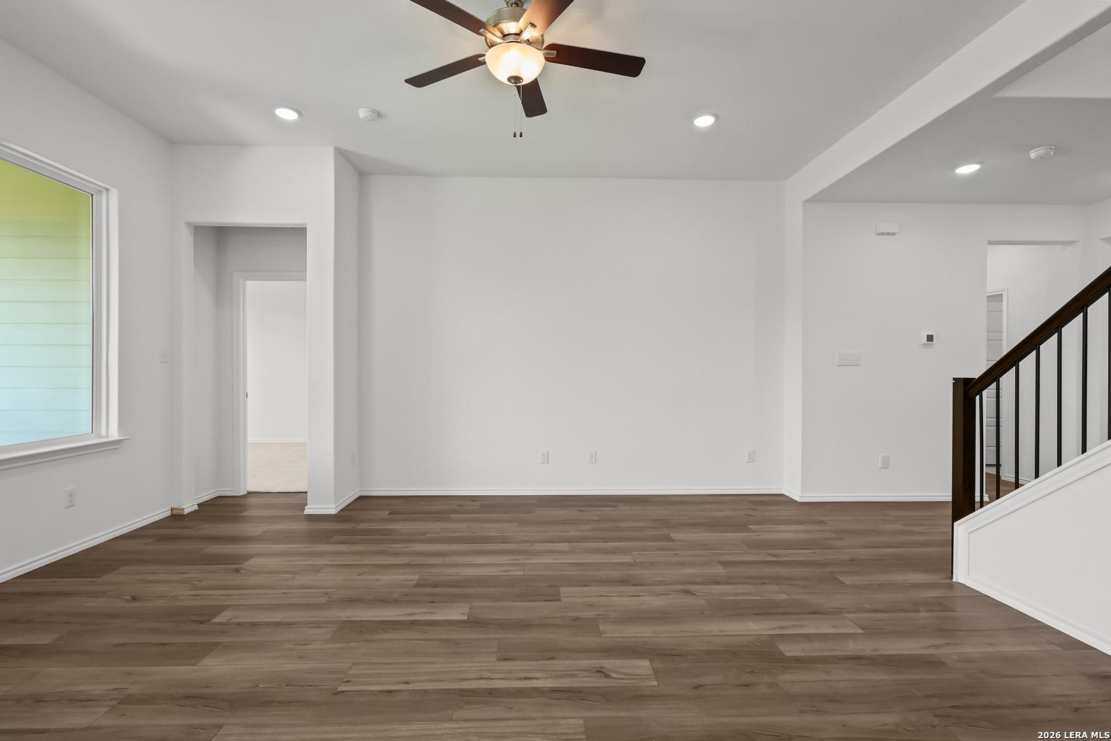Spacious empty living room with hardwood floors, ceiling fan, large window, and staircase in Davidson Homes The Sequoia B, San Antonio