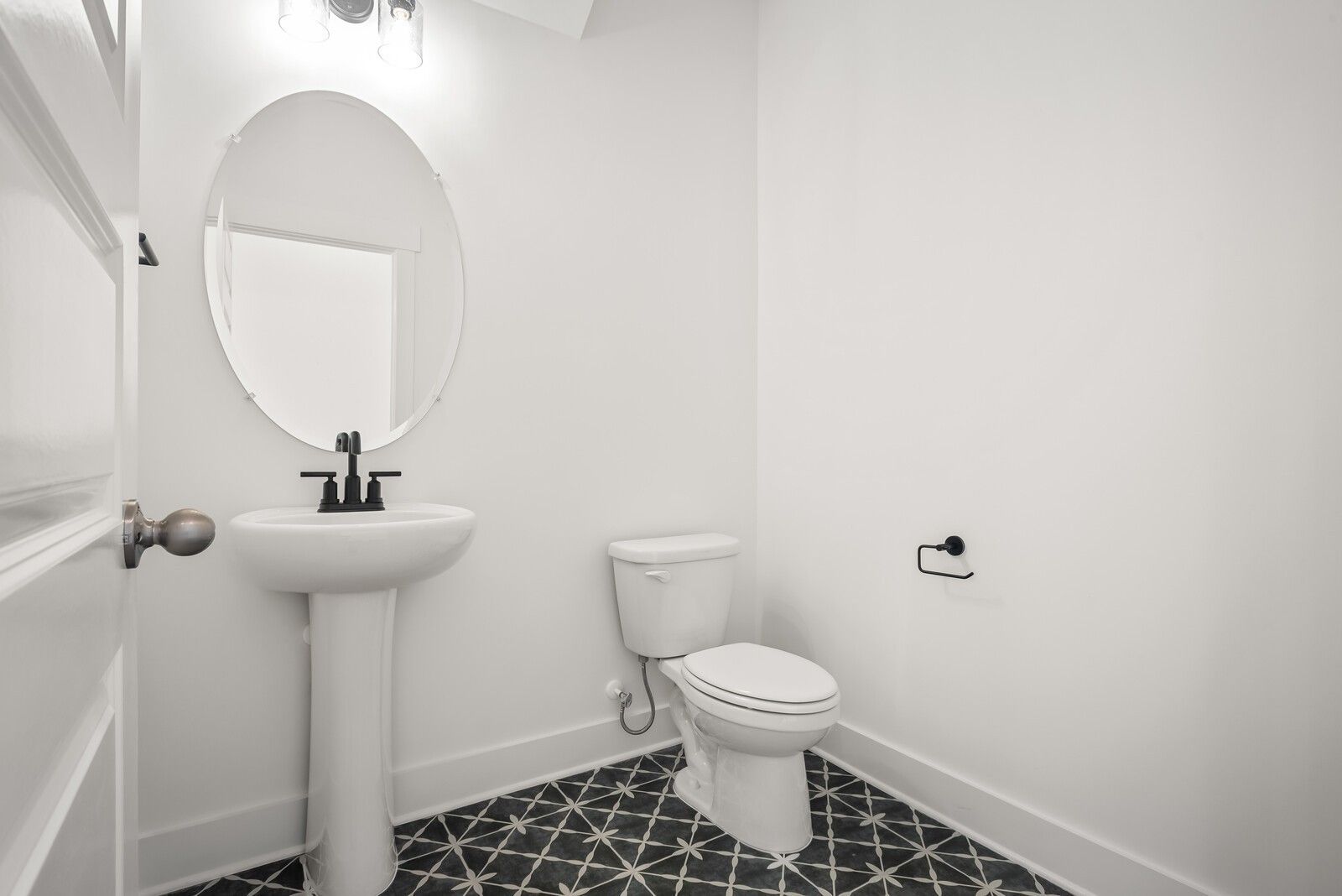 Elegant powder room with white pedestal sink, oval mirror, toilet, and geometric black-white tile floor in Davidson Homes The Logan C, Gallatin, TN