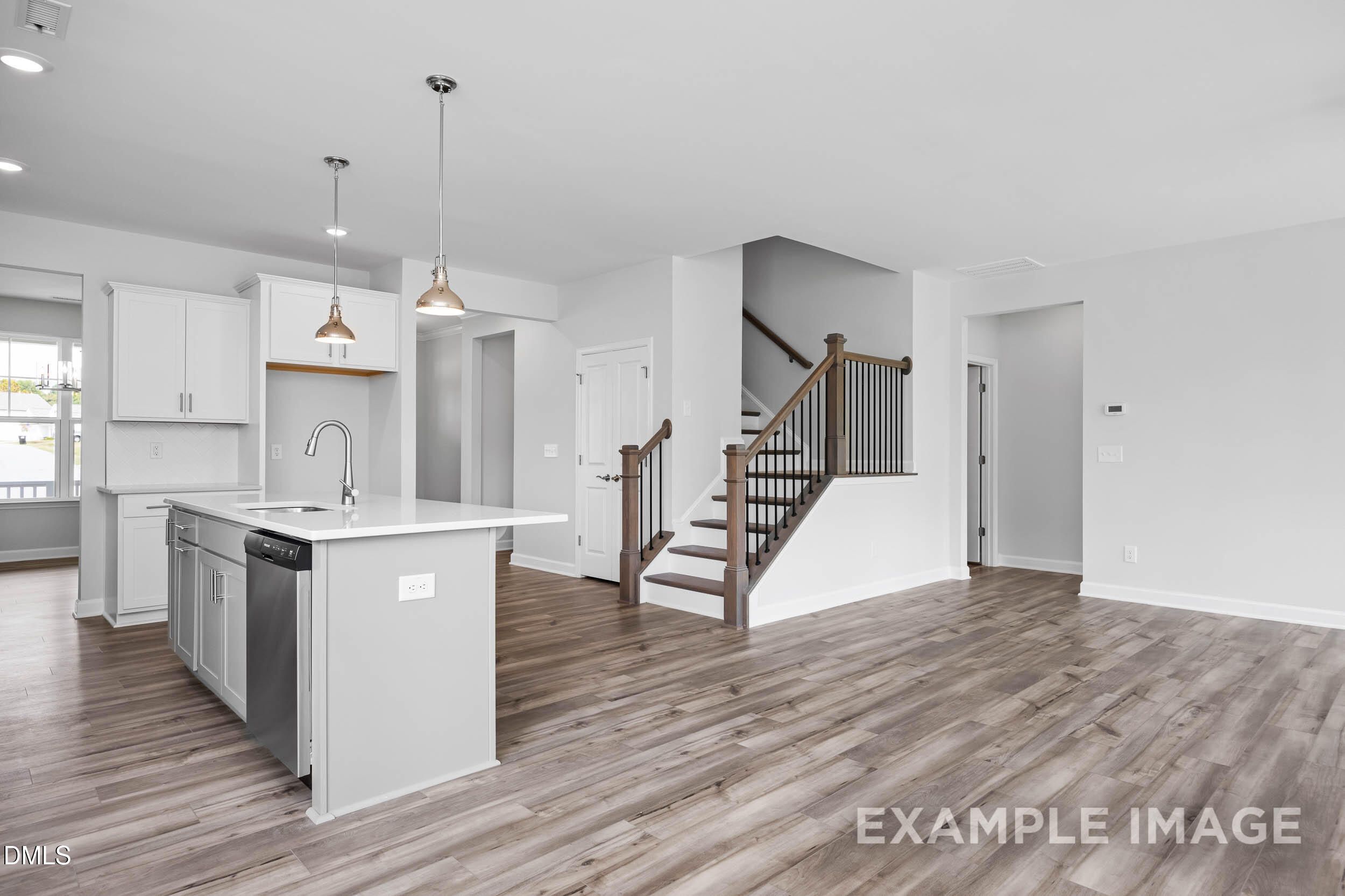 Modern open-concept kitchen with white cabinets, large island sink, and hardwood floors leading to wooden staircase in Davidson Homes The Ash B, Lillington, NC