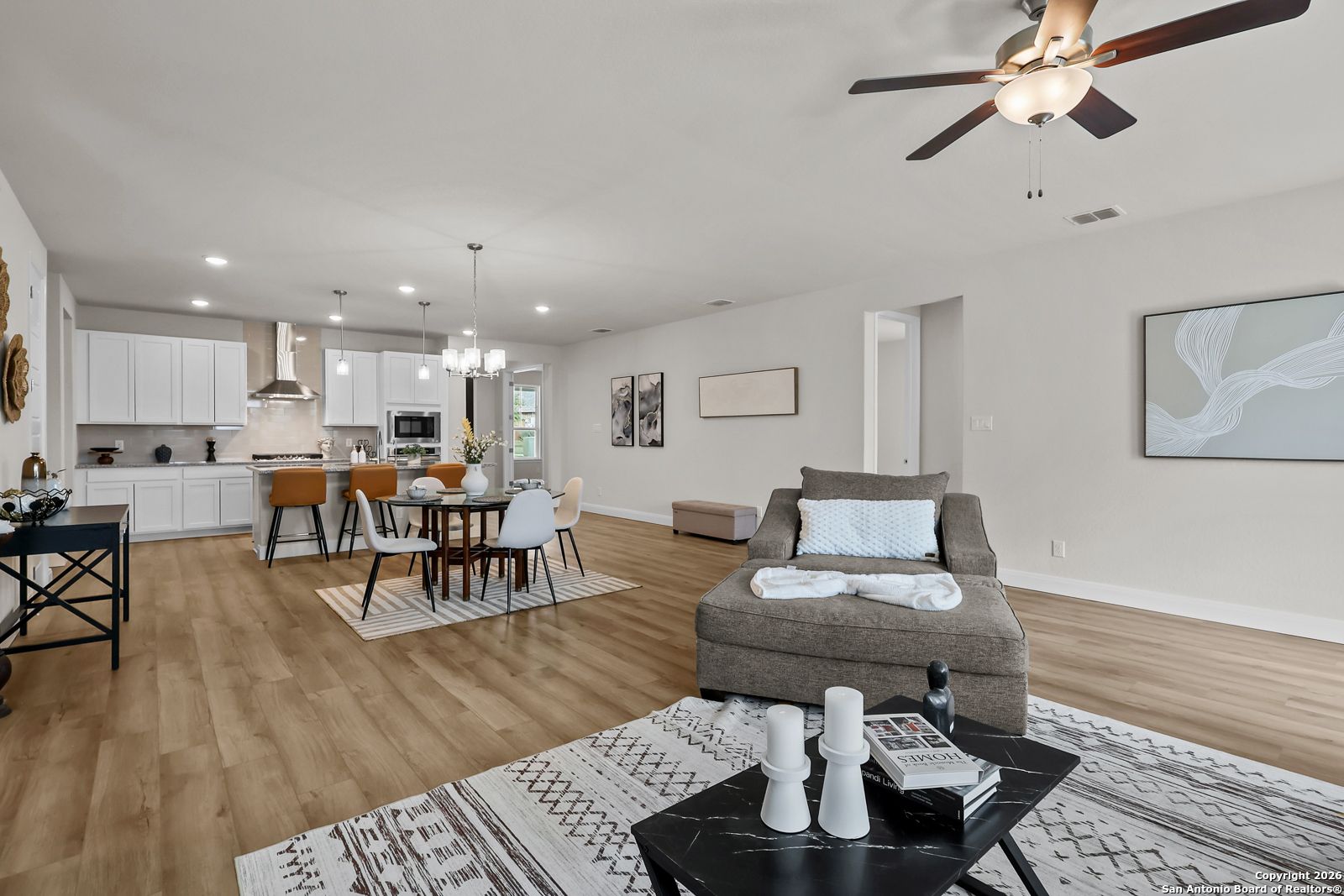 Open-concept living and kitchen with white cabinets, island bar, dining area, and plush gray sofa in The Lanier H, Ladera, San Antonio