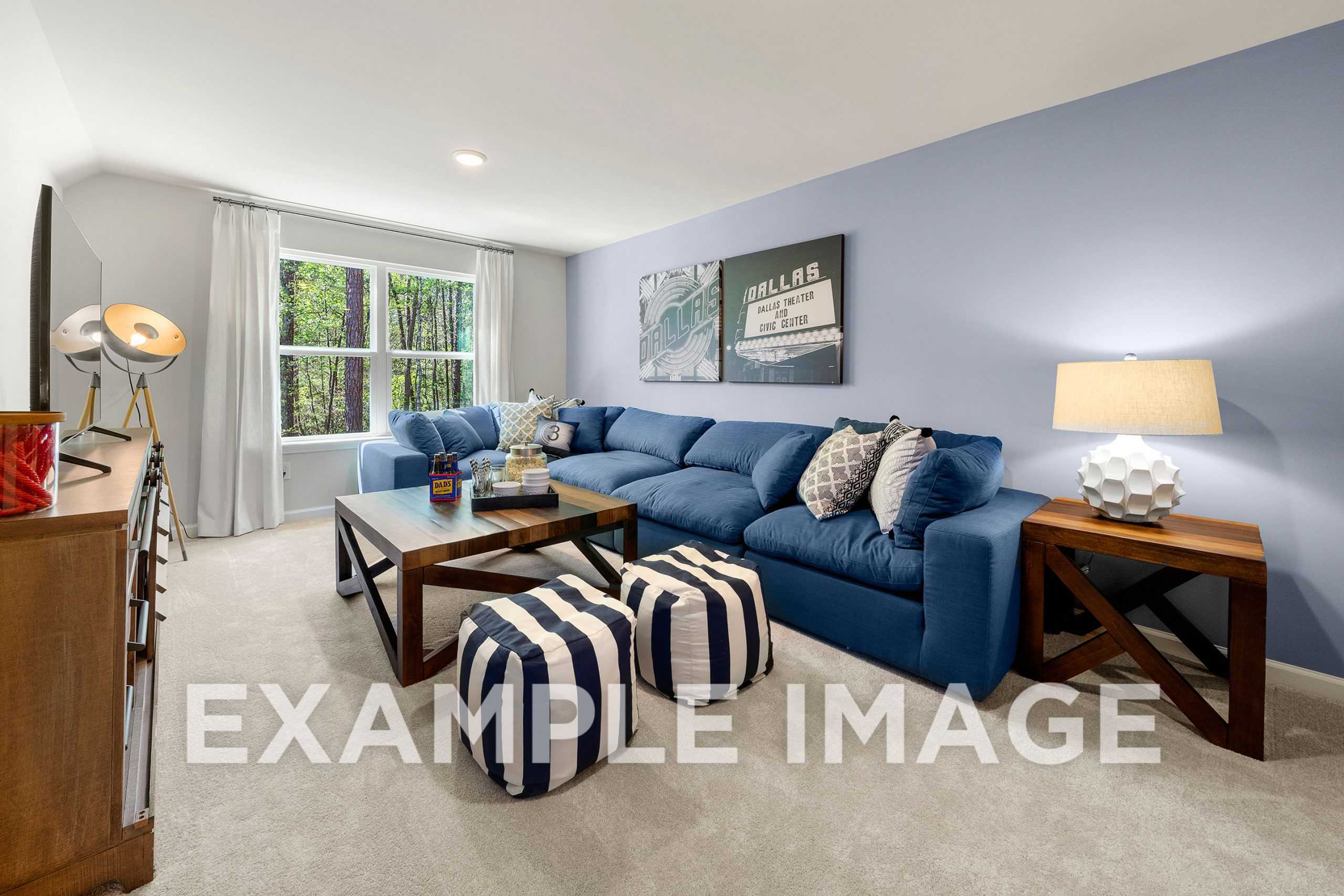 Cozy bonus room in The Hickory A featuring blue L-shaped sectional sofa, wooden coffee table, striped poufs, and large window with tree views