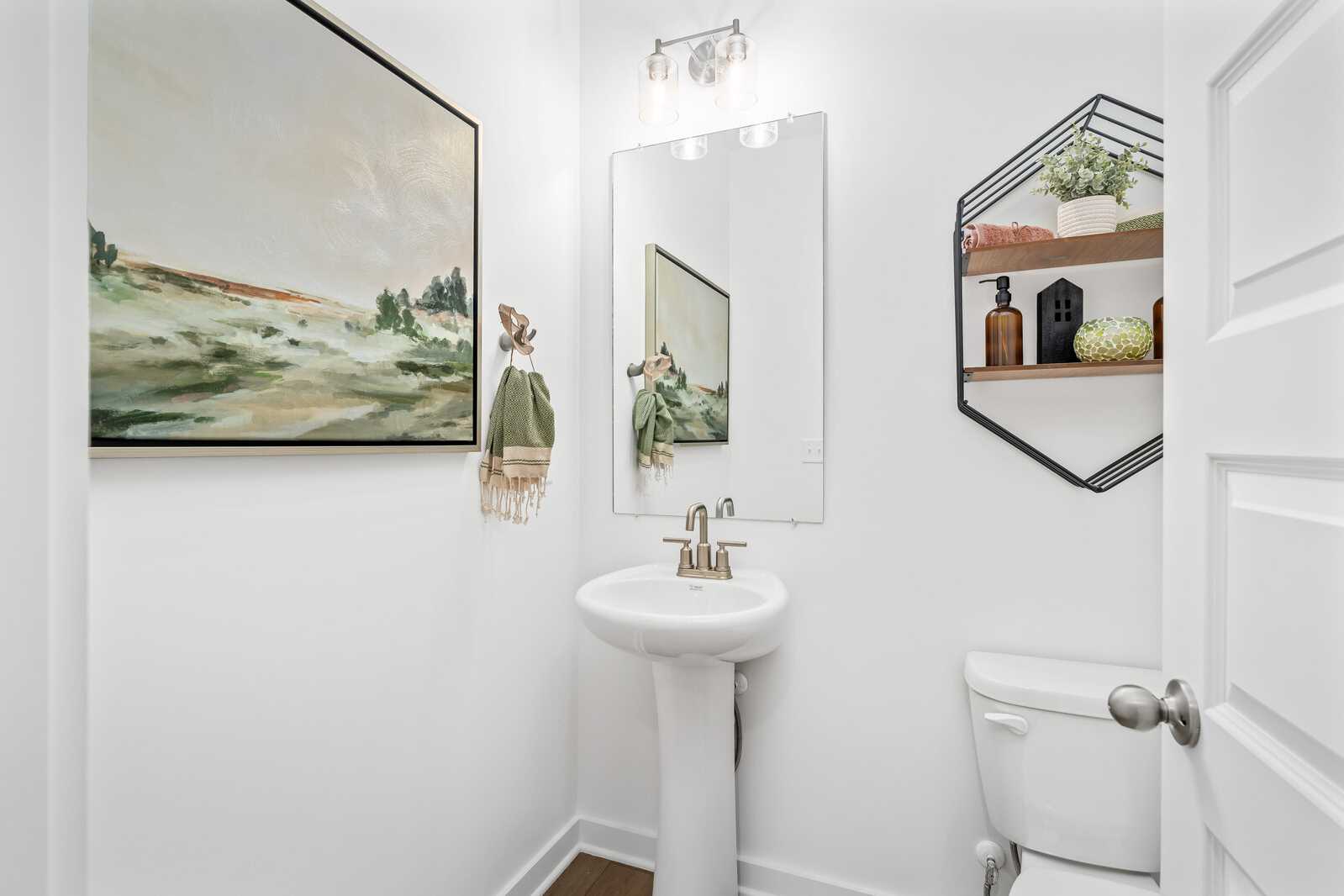 Bright white powder room in Calista Farms, White House TN with pedestal sink, round mirror, abstract landscape art, and hexagonal shelves