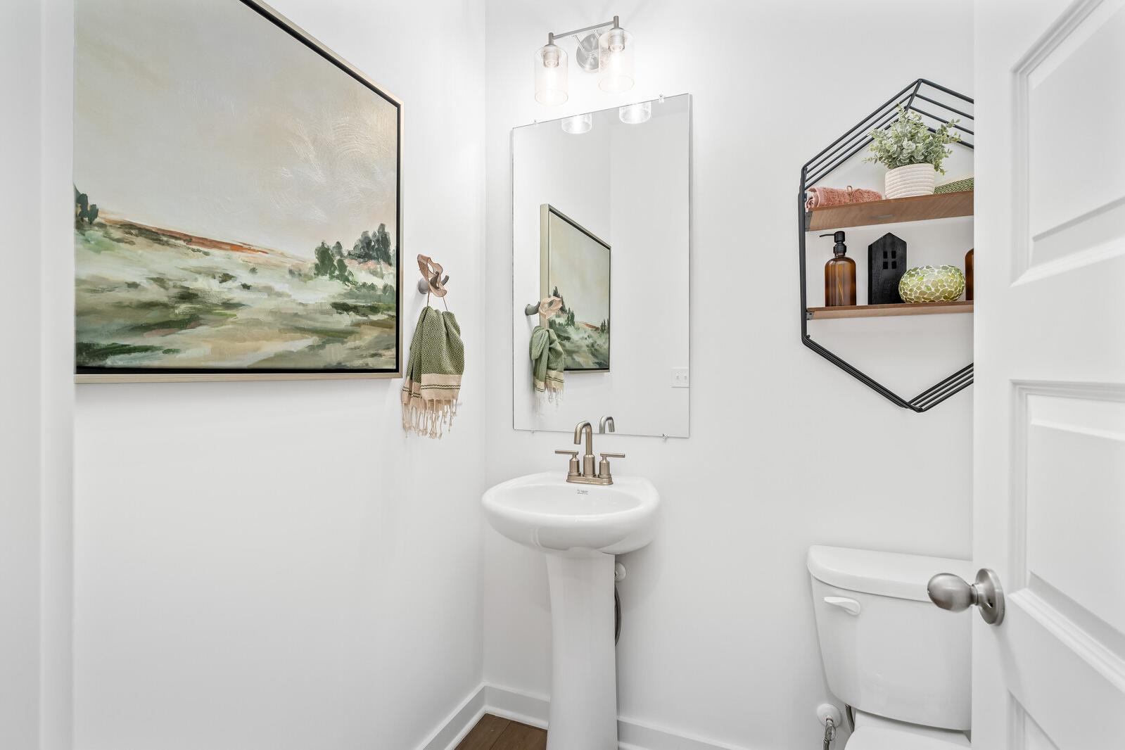 Bright white powder room in Calista Farms, White House TN with pedestal sink, round mirror, abstract landscape art, and hexagonal shelves
