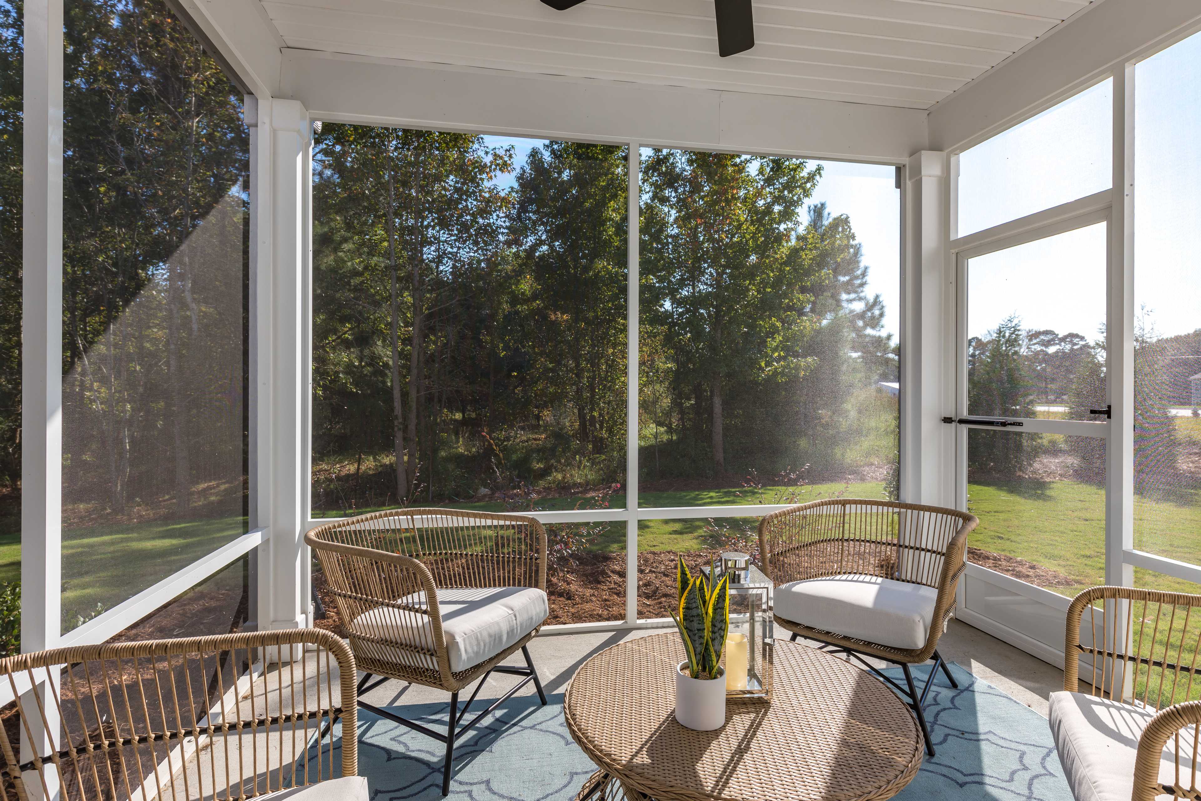 Screened porch with rattan chairs, coffee table, plants and ceiling fan at Enclave at Belmont in Belmont NC overlooking wooded yard