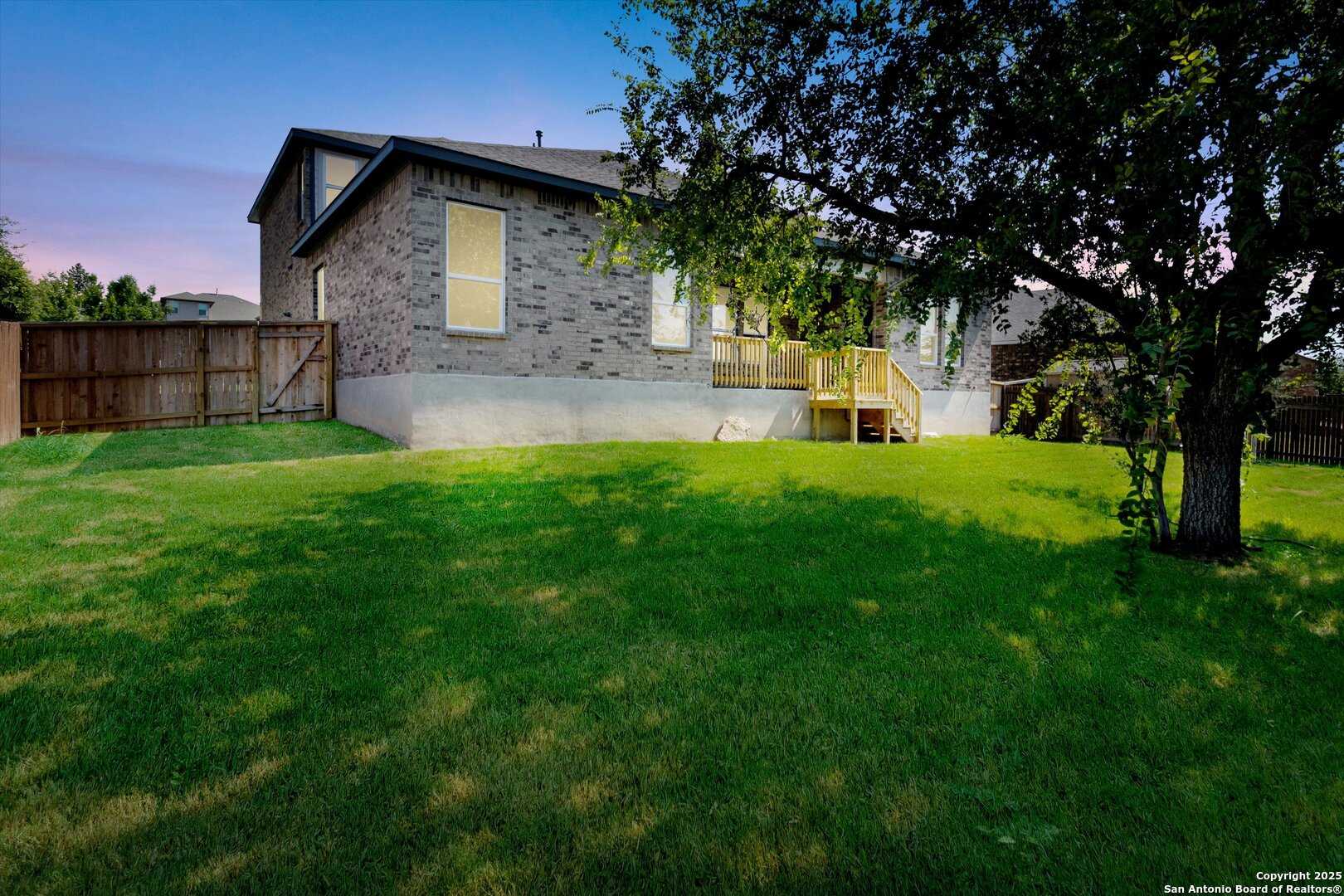 Backyard view of two-story brick home with wooden deck, large windows, lush green lawn, and mature apple tree in Ladera, San Antonio, Texas