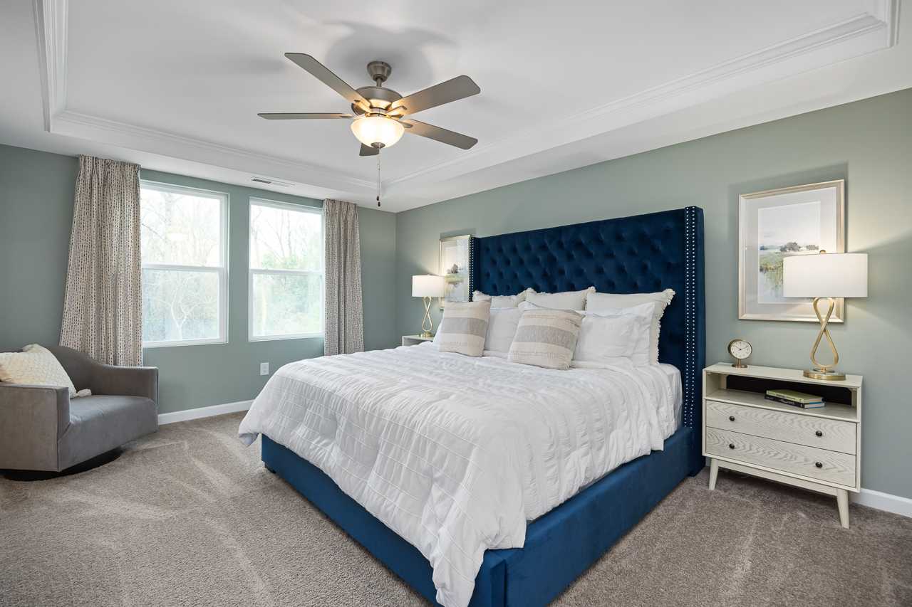 Spacious primary bedroom at Gregory Village Townhomes in Lillington NC with navy velvet headboard, white bedding, and ceiling fan