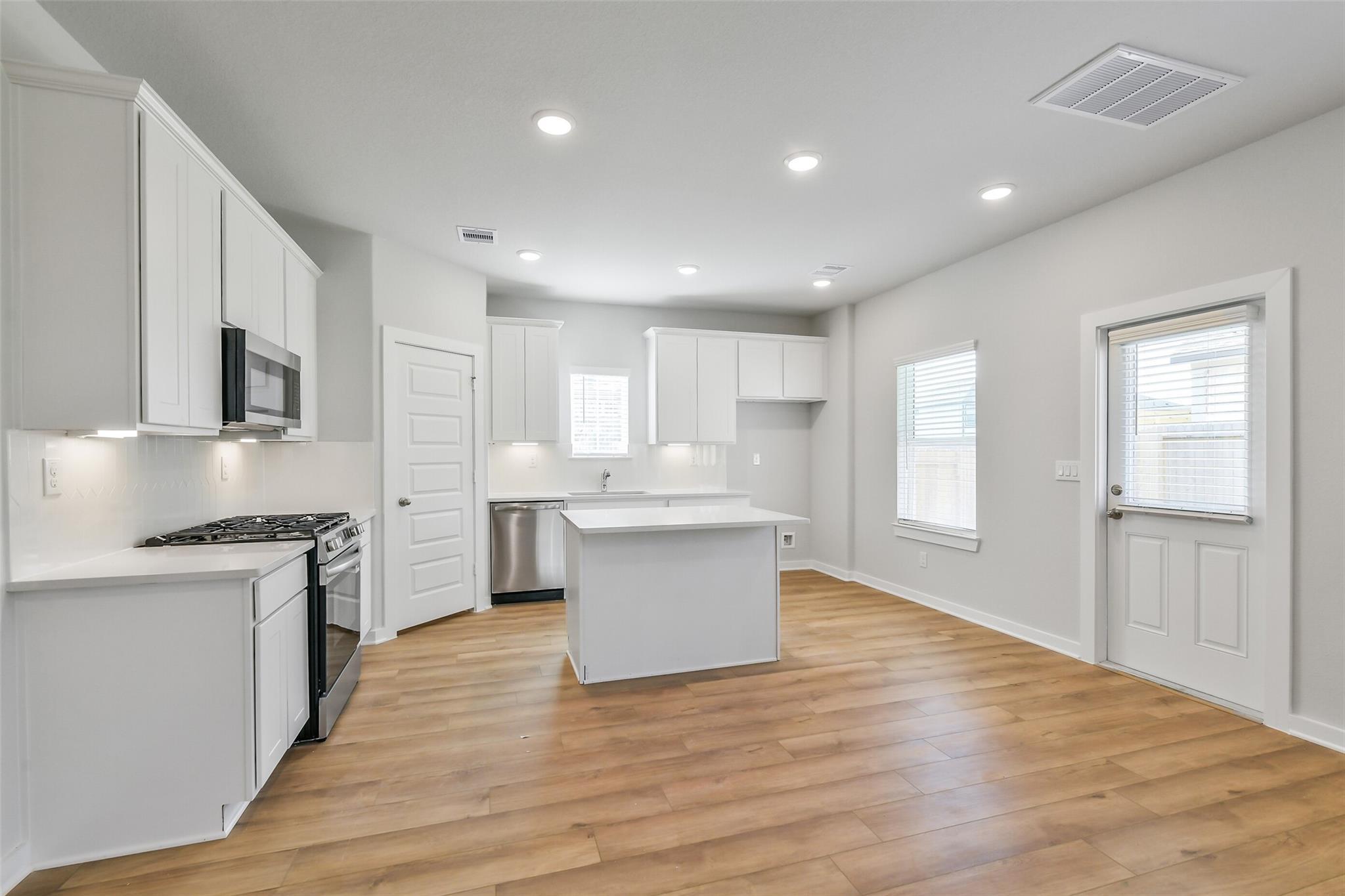 Modern white kitchen with center island, stainless steel appliances, and hardwood floors in Davidson Homes The Trinity F, Magnolia, Texas