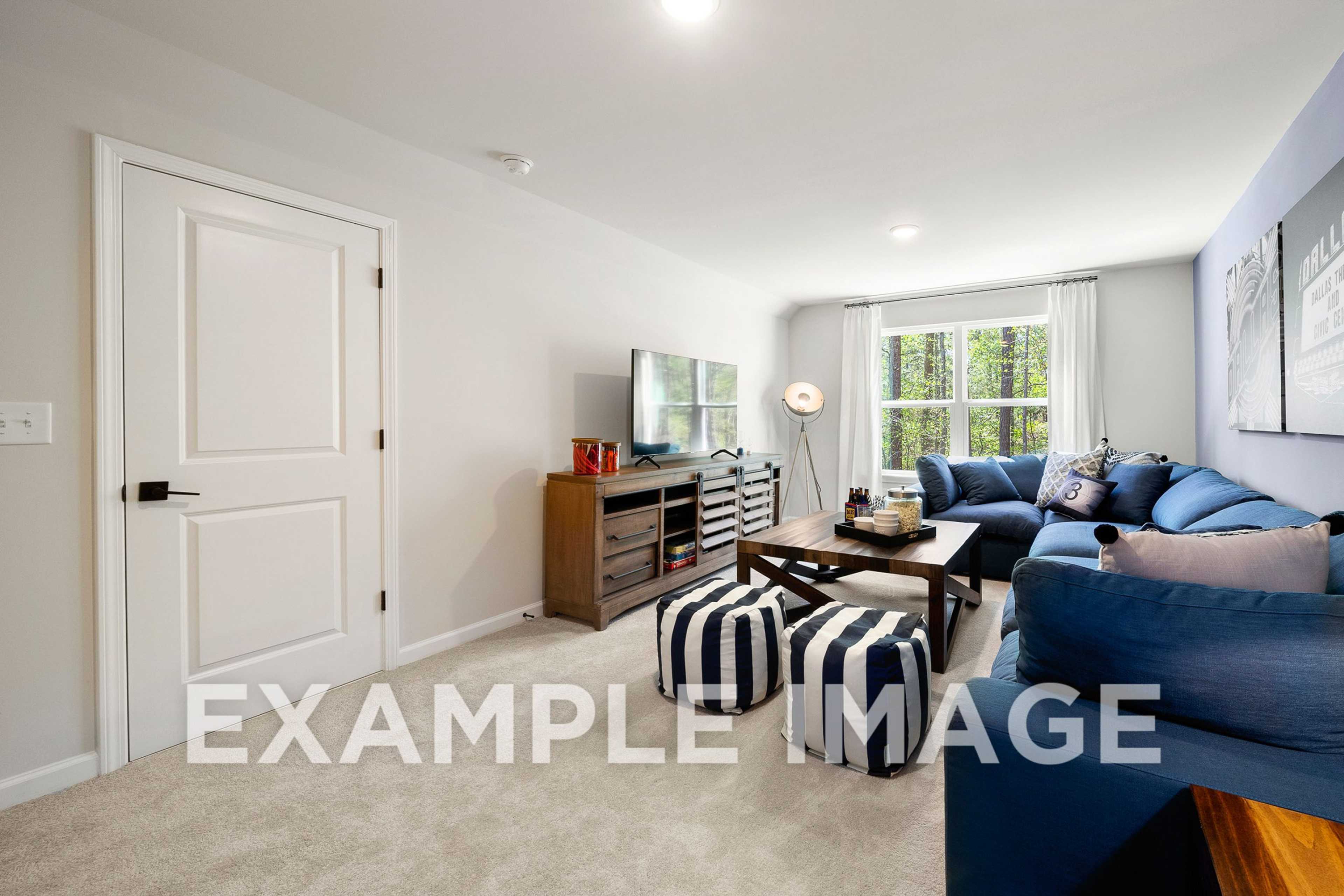 Cozy bonus room in The Hickory A home with navy blue sectional sofa, wooden TV console, striped poufs, and large window overlooking trees