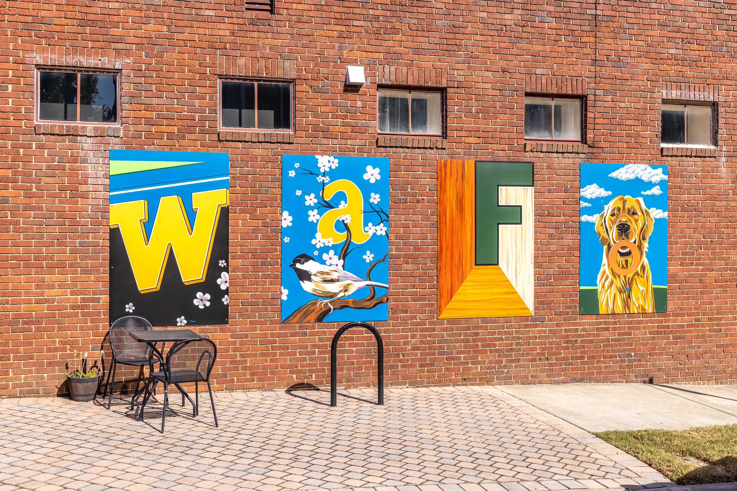 Vibrant brick wall mural at Sage on North Main in Wake Forest NC featuring colorful WAFE letters with bird, flowers, door, golden retriever, bistro seating