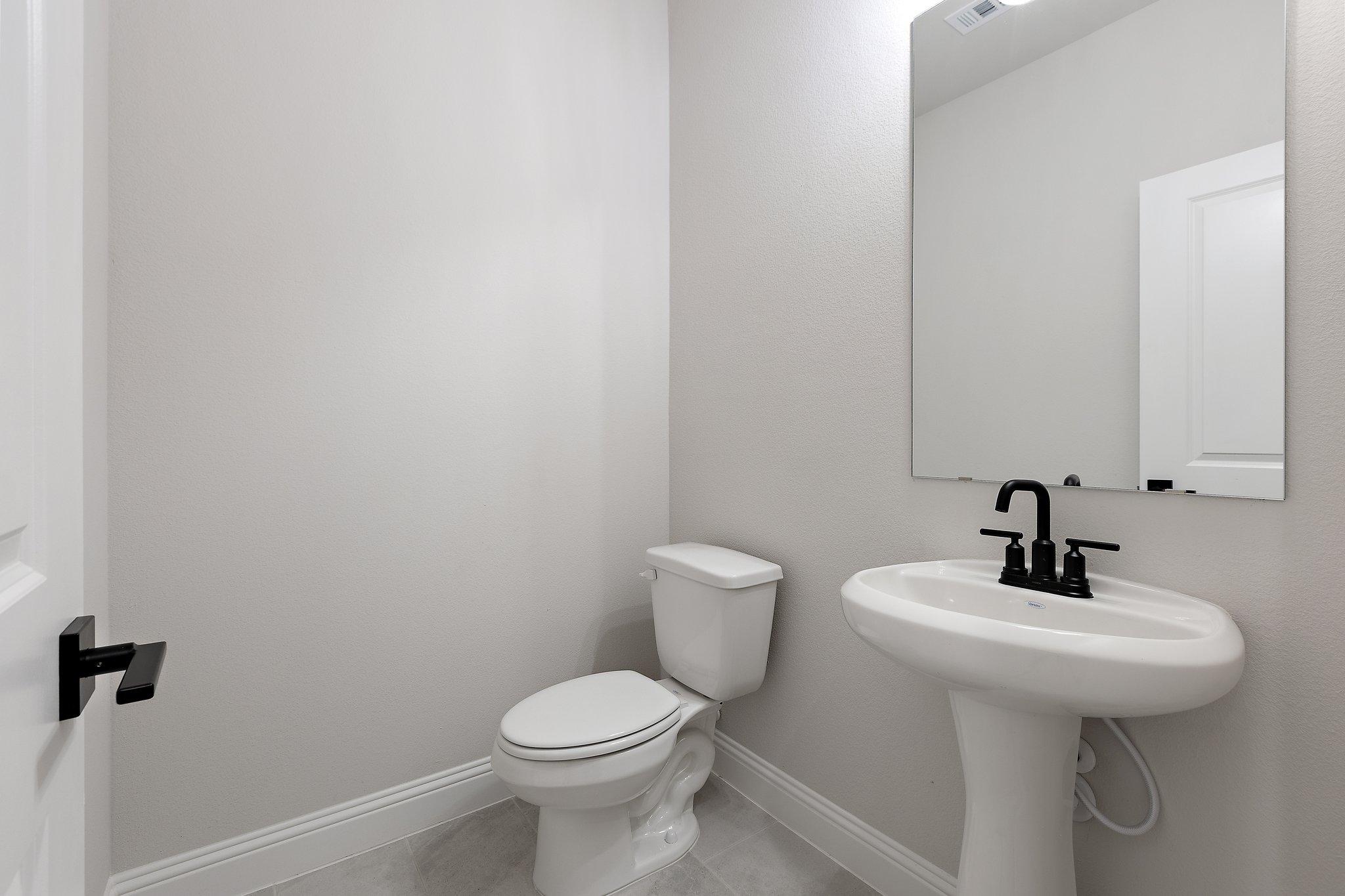 Spacious powder room in The Harrison D featuring white pedestal sink, black faucet, large mirror, and toilet