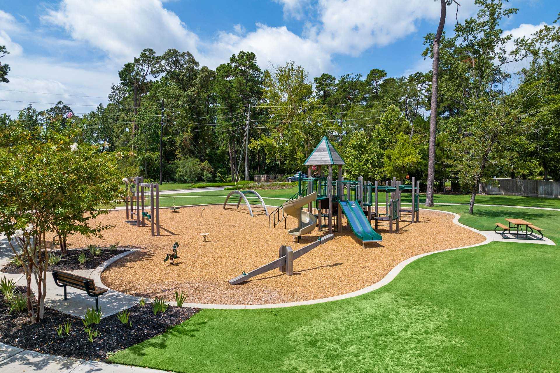 Children's playground at Lakes at Black Oak in Magnolia Texas with slides climbers swings sand pit benches and picnic table amid trees