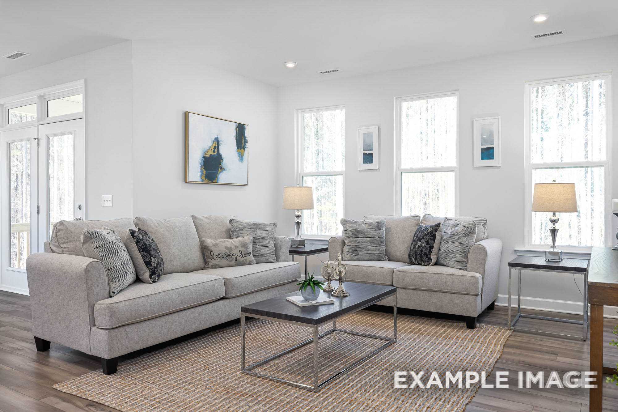 Cozy living room in The Ash C home design with light gray sofas, coffee table, lamps, abstract art, and large windows
