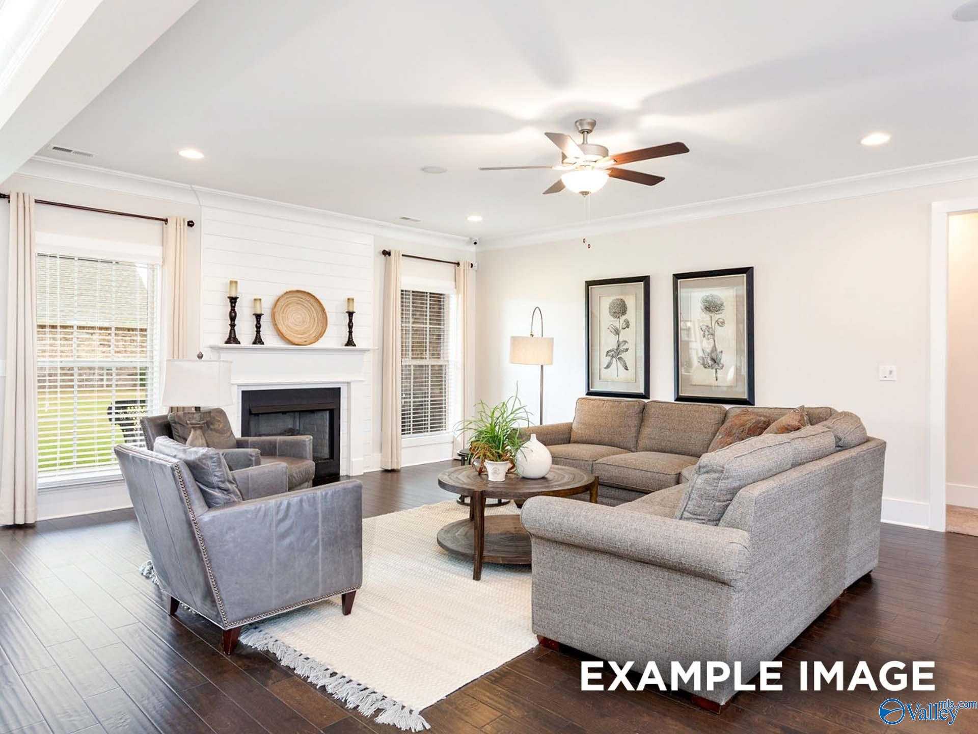 Cozy living room with beige L-shaped sofa, gray armchair, wood fireplace, ceiling fan, large windows in Davidson Homes The Finleigh, Meridianville, Alabama