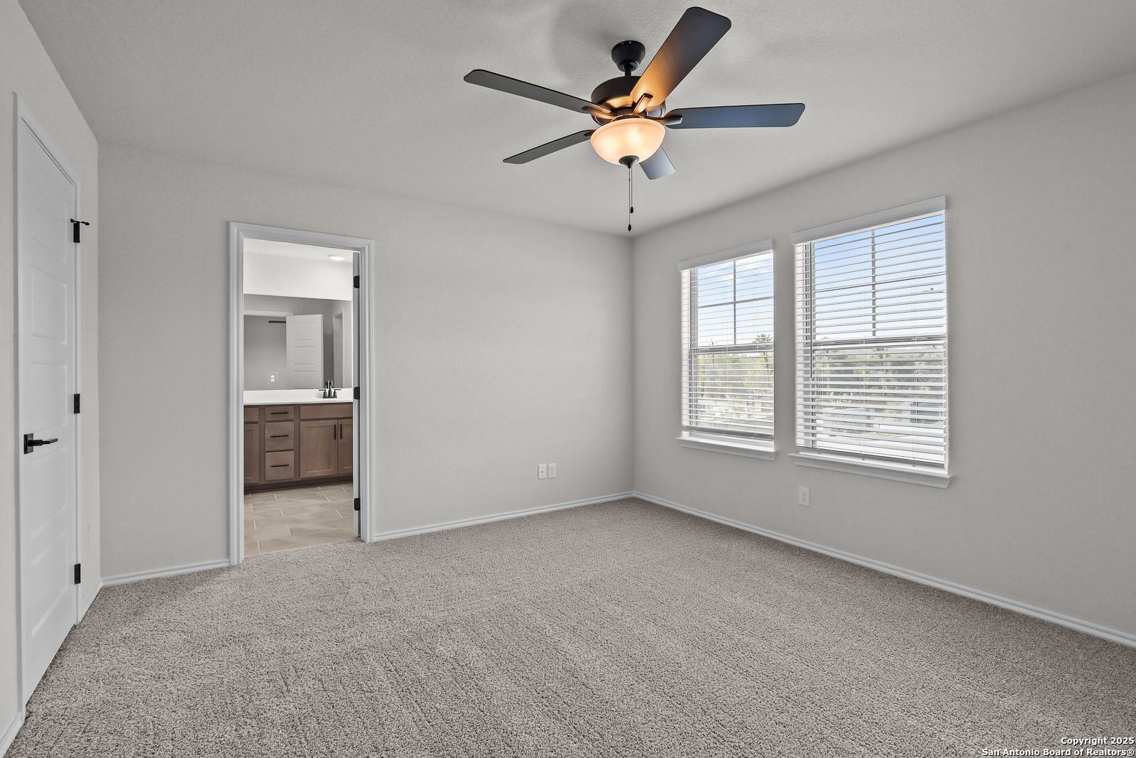 Spacious bedroom with ceiling fan, large windows, blinds, and adjacent bathroom in Davidson Homes The Florence C, San Antonio
