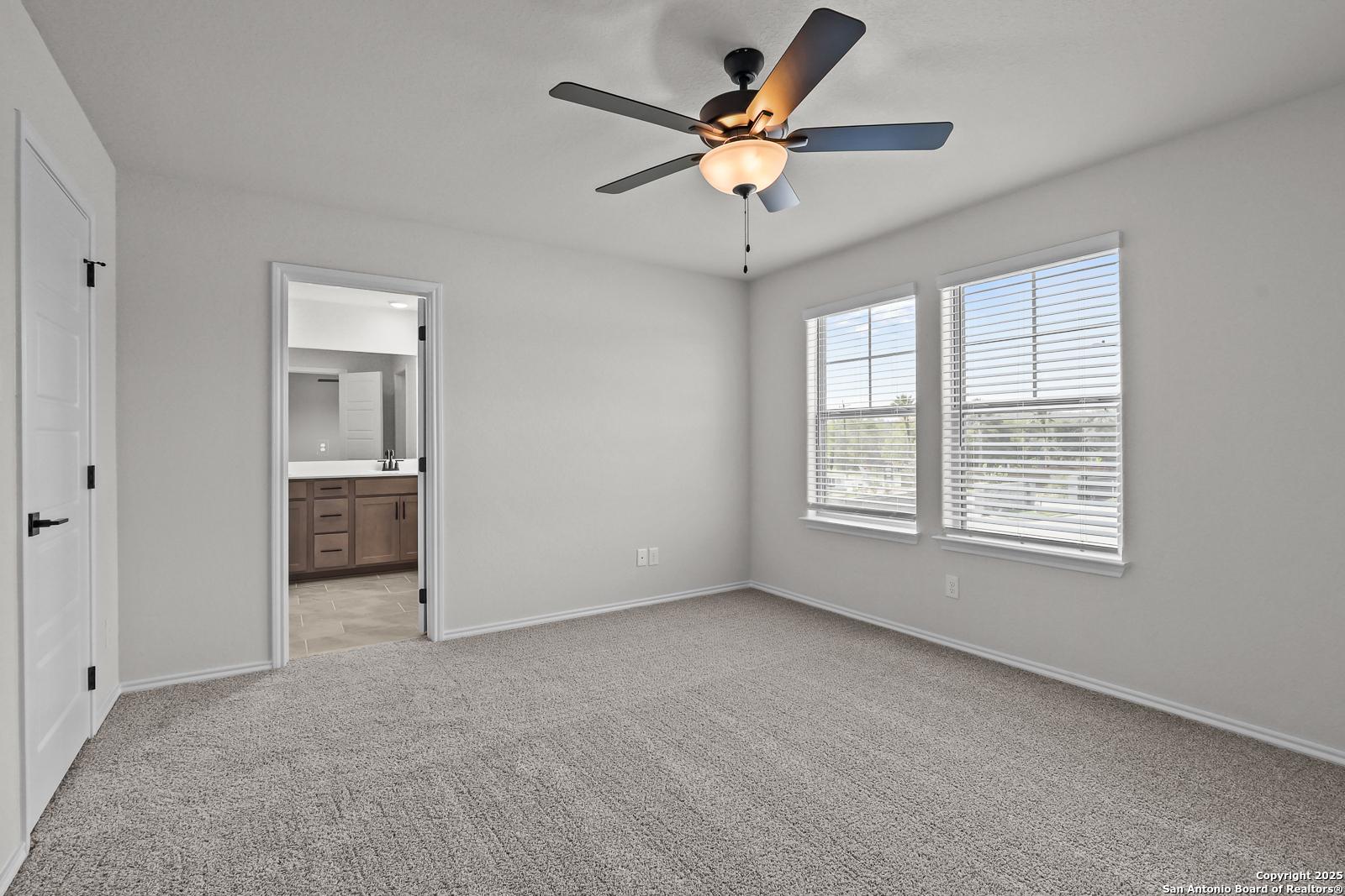Spacious bedroom with ceiling fan, large windows, blinds, and adjacent bathroom in Davidson Homes The Florence C, San Antonio