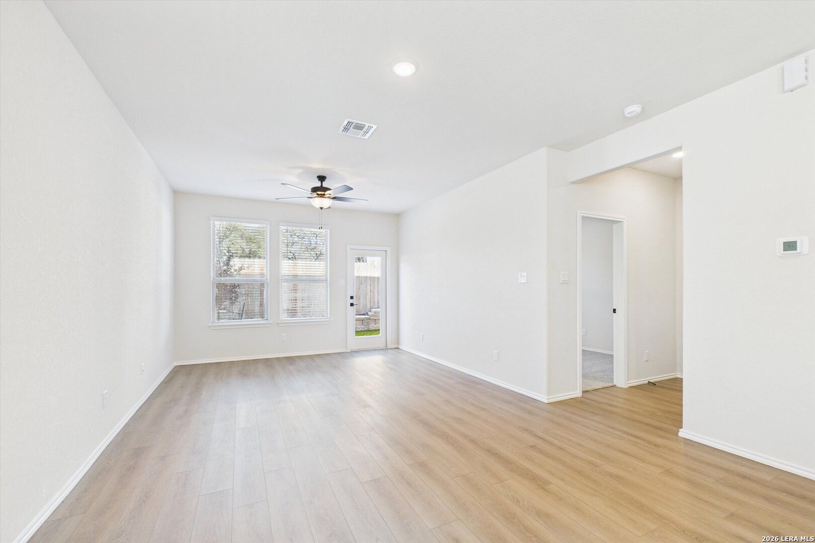 Open living area with light hardwood floors, ceiling fan, large windows, and glass door to backyard in Davidson Homes The Gillian B, San Antonio