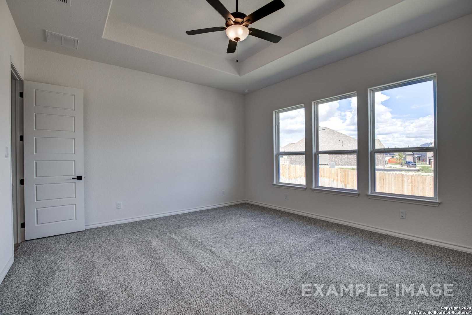 Bright bedroom with tray ceiling, ceiling fan, gray carpet, and large windows to backyard in Davidson Homes The Garner C, Castroville, Texas