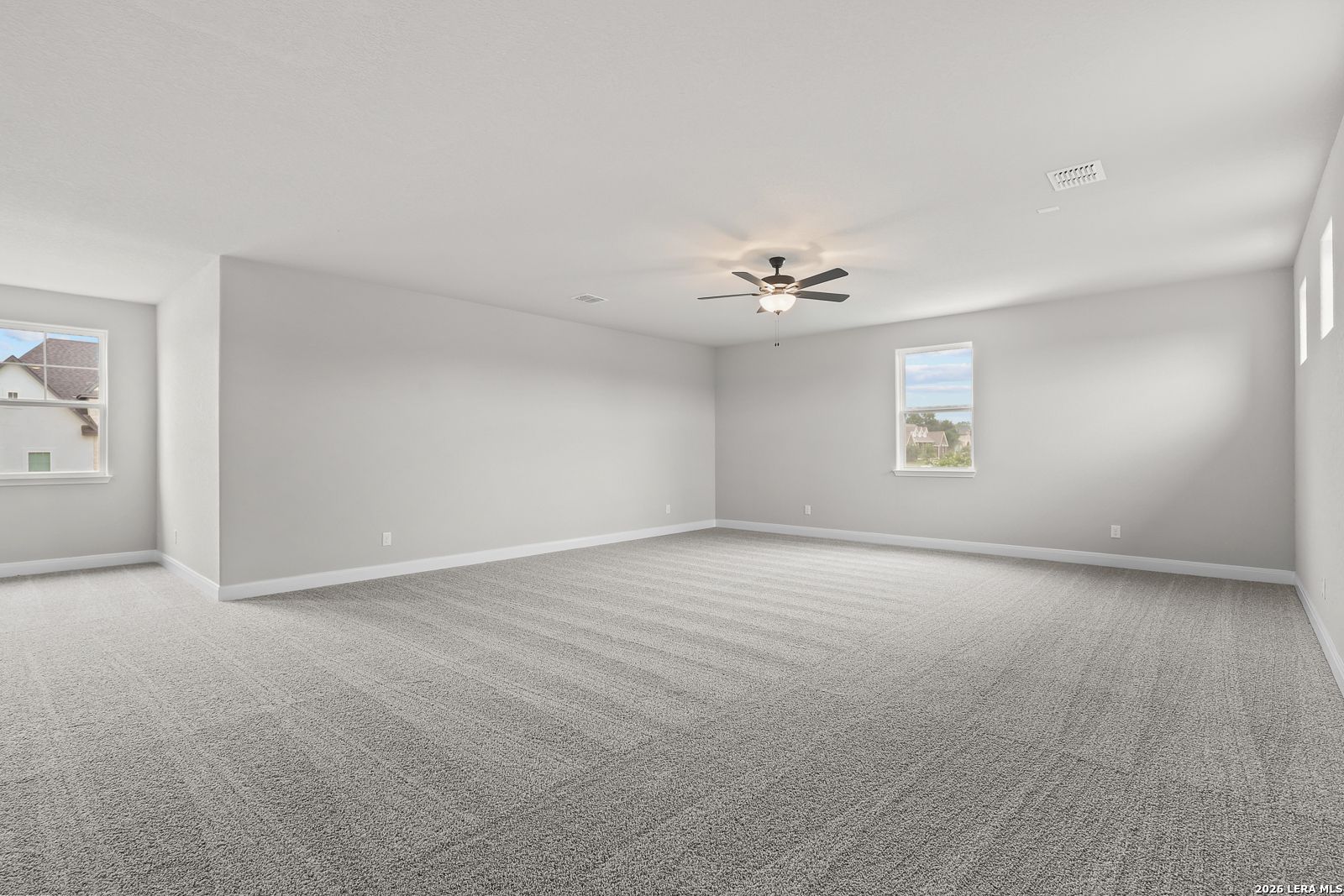 Spacious empty bedroom with gray carpet, neutral walls, ceiling fan, and windows overlooking neighborhood in Davidson Homes The Belmont H, Castroville, Texas