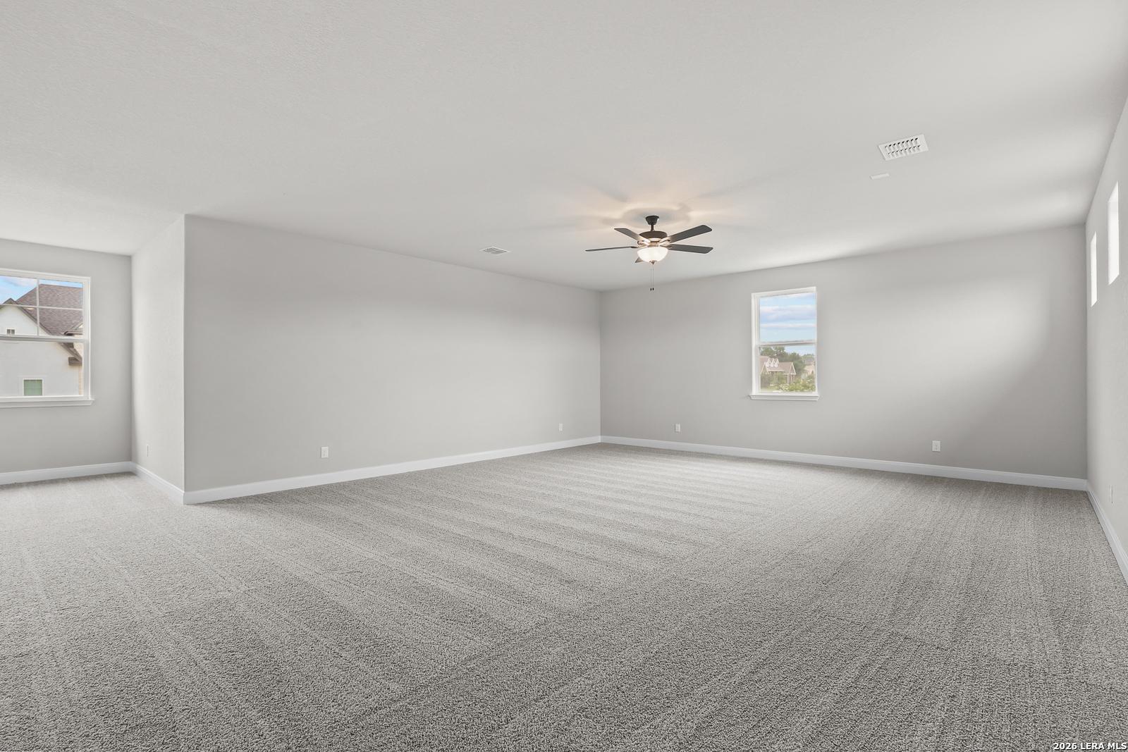 Spacious empty bedroom with gray carpet, neutral walls, ceiling fan, and windows overlooking neighborhood in Davidson Homes The Belmont H, Castroville, Texas