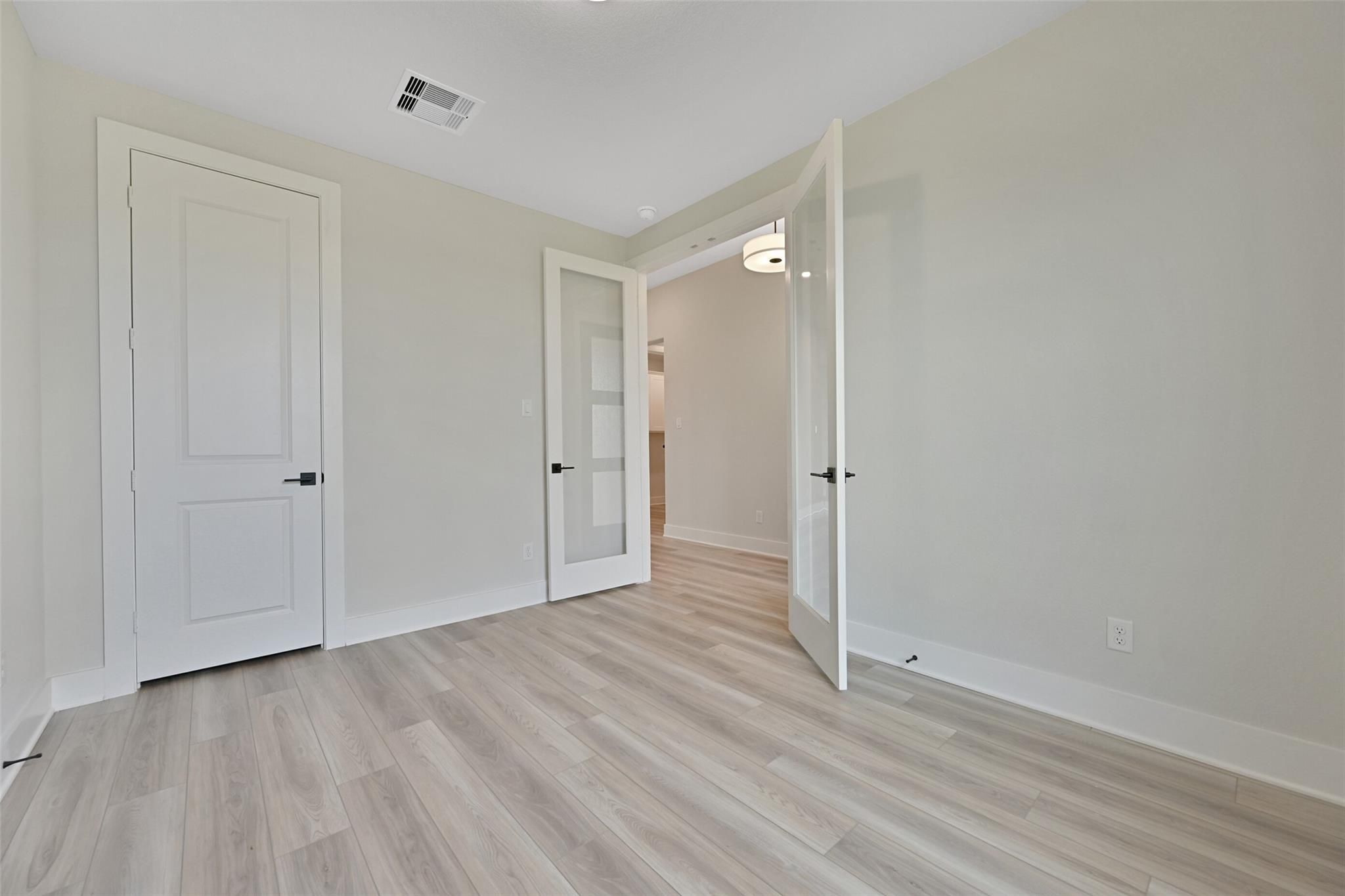 Spacious bedroom with light wood floors, beige walls, and open glass door to en-suite bath in Davidson Homes The Edward C, Lago Mar, Texas City