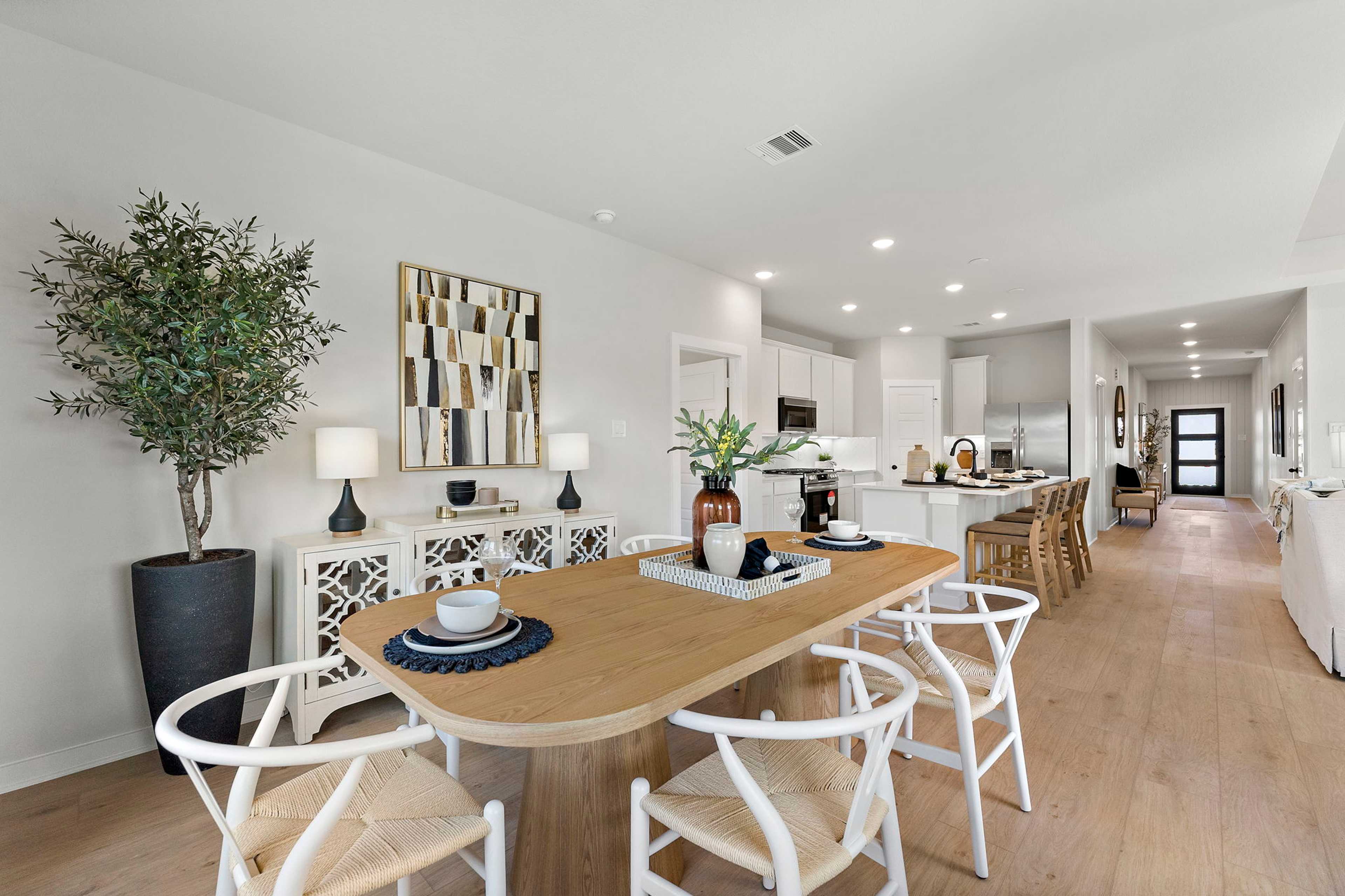 Open-concept dining area at Sundance Cove in Crosby Texas with oak table, white woven chairs, potted olive tree, and adjacent white kitchen