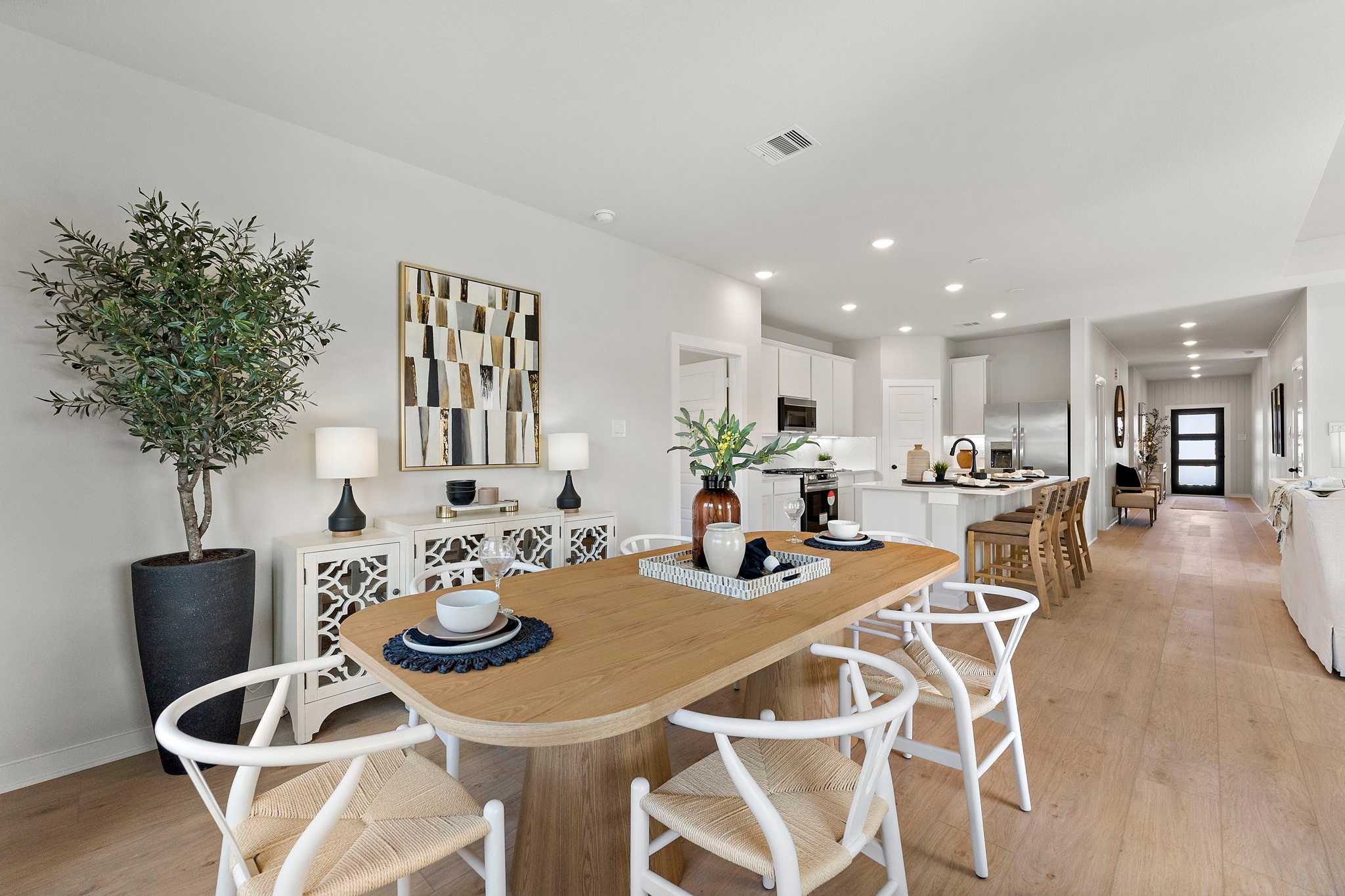 Open-concept dining room in Sundance Cove Crosby Texas by Davidson Homes with light wood table, white rattan chairs, potted olive tree, and modern kitchen