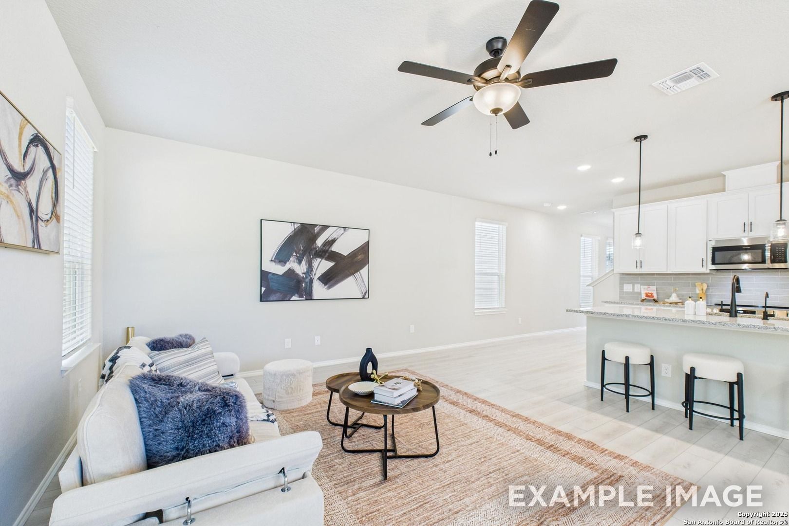 Open-concept living room with white sectional sofa, abstract art, and ceiling fan adjacent to white kitchen cabinets in Davidson Homes The Charlotte A, San Antonio