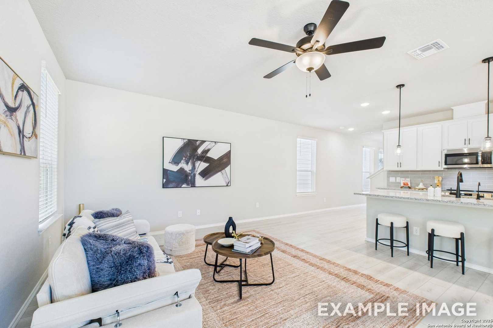 Open-concept living room with white sectional sofa, abstract art, and ceiling fan adjacent to white kitchen cabinets in Davidson Homes The Charlotte A, San Antonio