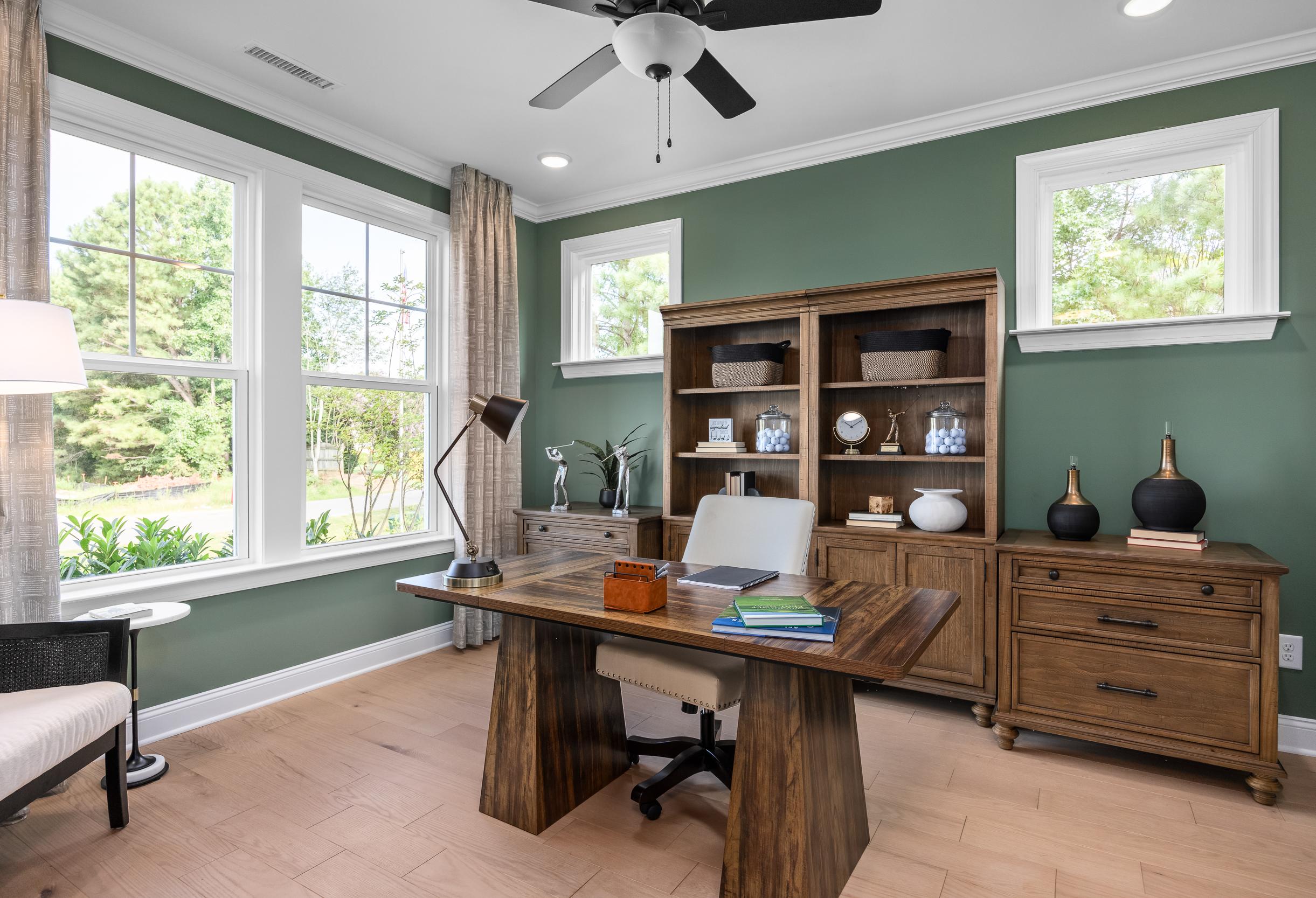 Spacious study in The Crawford home design featuring wooden desk, built-in bookshelves, green walls, and large windows with tree views