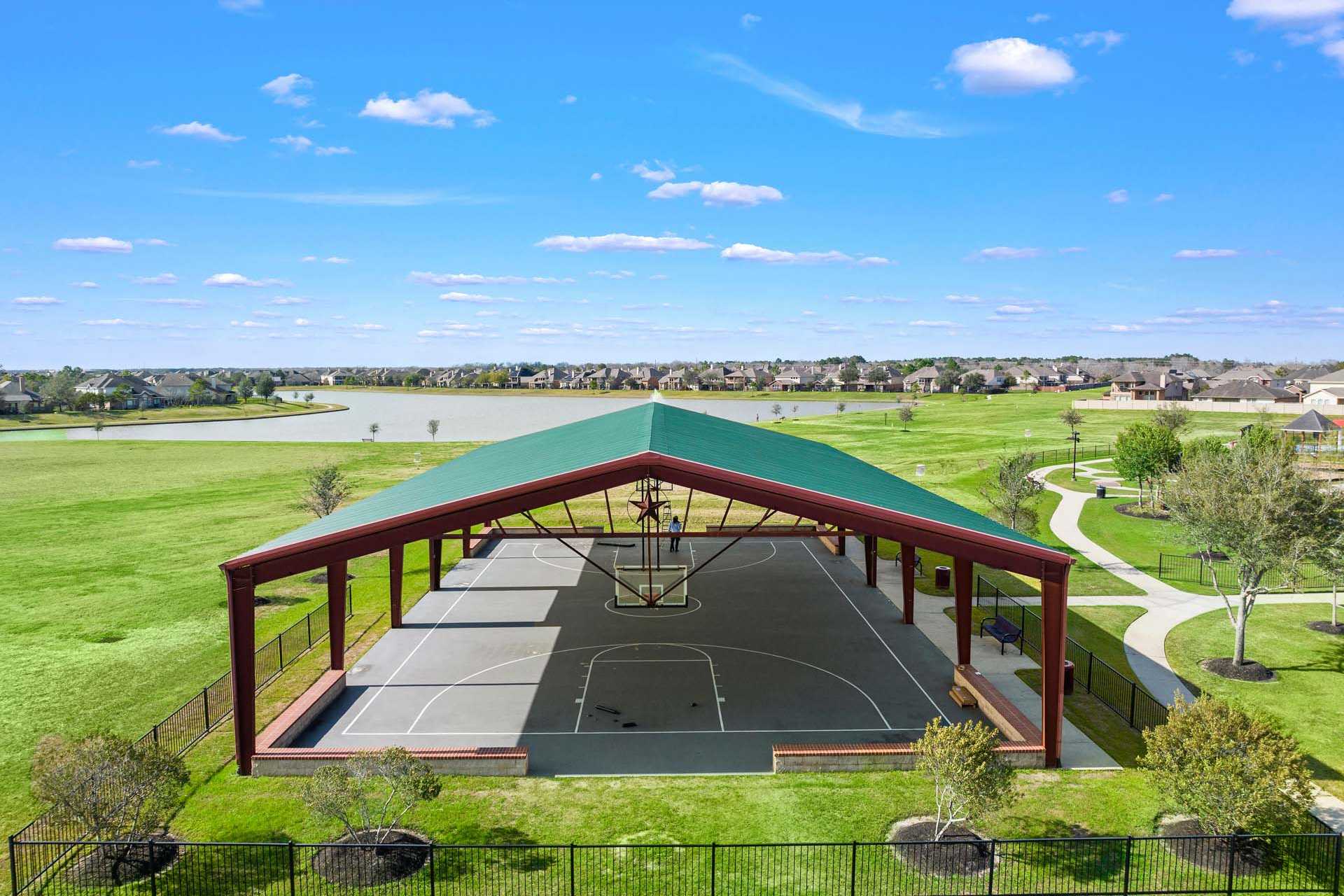 Covered basketball court pavilion with green roof at Sierra Vista in Rosharon, Texas surrounded by grassy fields, trees, and lake