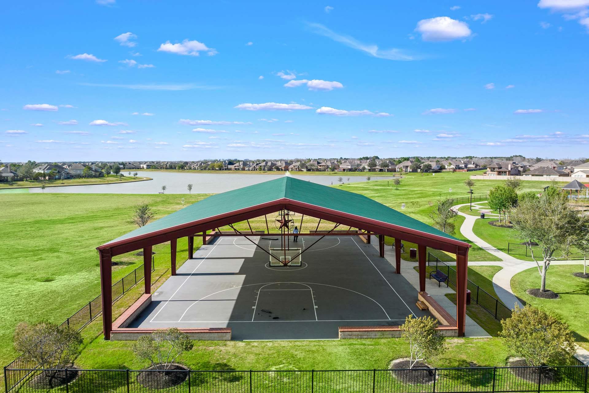 Covered basketball court pavilion with green roof at Sierra Vista in Rosharon, Texas surrounded by grassy fields, trees, and lake