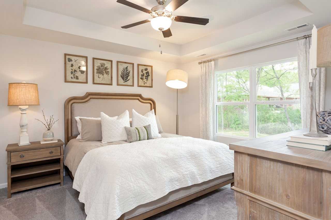 The Franklin master bedroom with tufted beige headboard bed, white quilt, botanical wall art, ceiling fan, and large window views