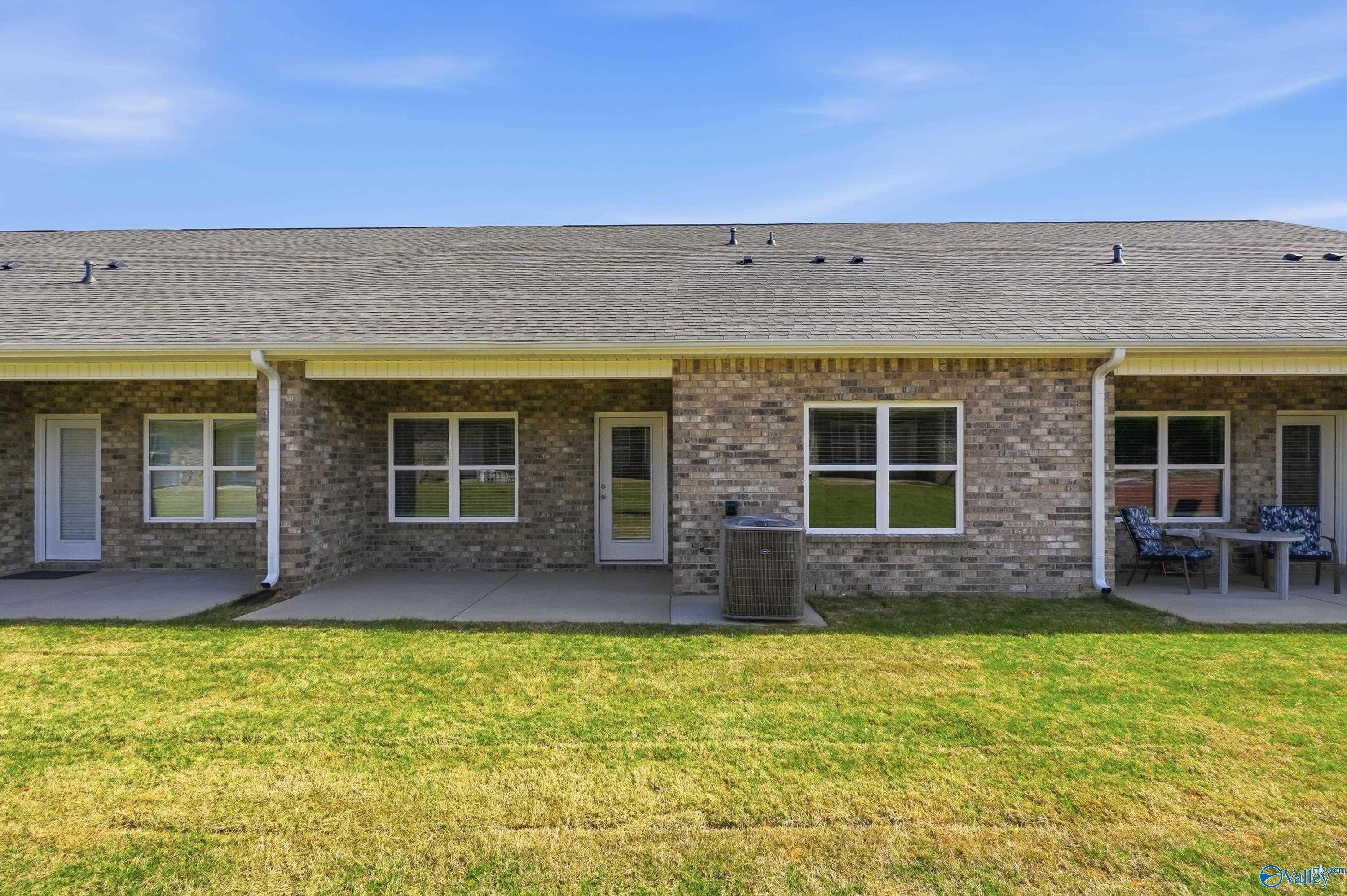 Rear brick exterior of single-story home with covered patio, outdoor seating, AC unit, and lush green yard in The Retreat at Hollon Meadow, Decatur, Alabama