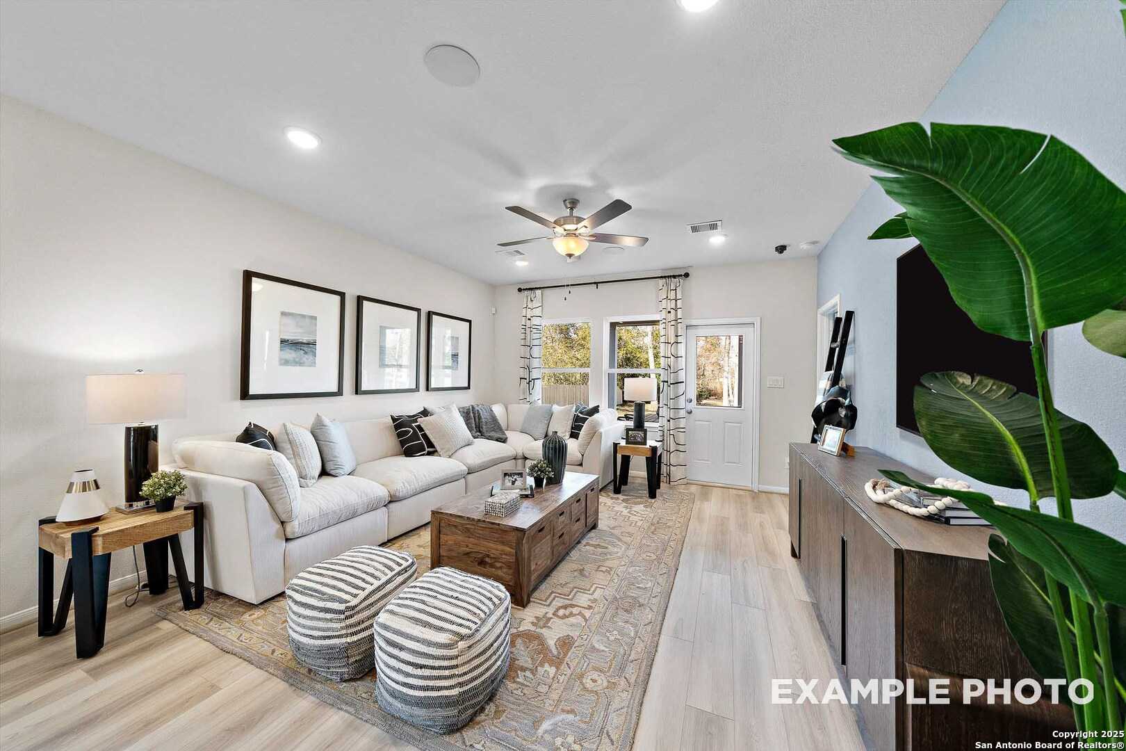 Modern living room with white L-shaped sofa, ceiling fan, large windows, and TV in The San Marcos D by Davidson Homes, San Antonio, Texas