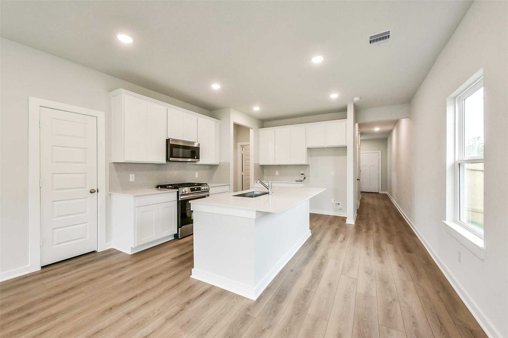 Modern white kitchen with large island, stainless steel appliances, and open hallway in Davidson Homes The Frio G, Dayton TX