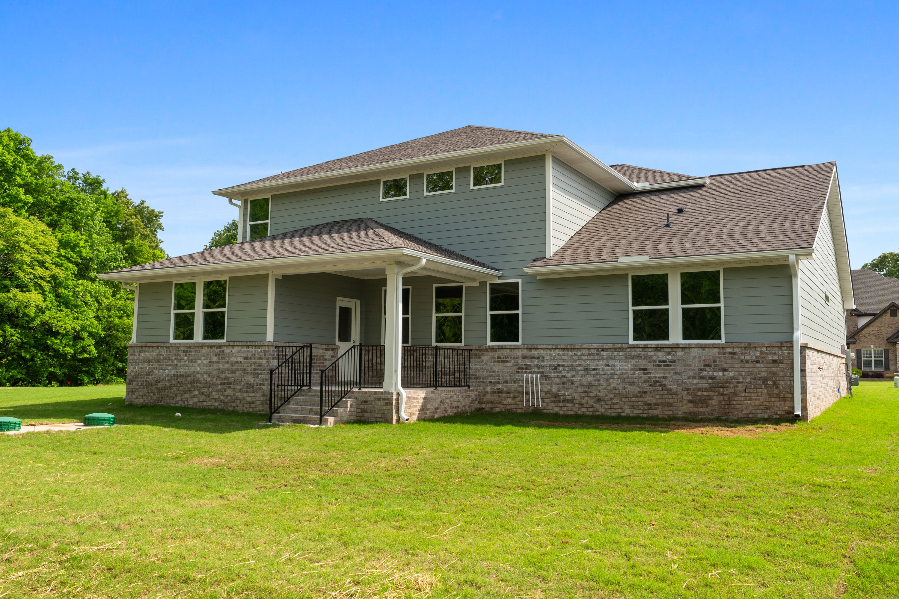 Rear elevation of The Avalon D two-story home featuring gray siding, brick base, covered porch, and lush green backyard in Owens Cross Roads, Alabama