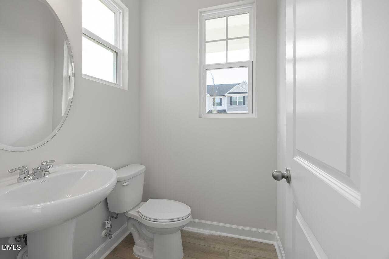 Modern powder room with white pedestal sink, round mirror, toilet, and window in Davidson Homes The Graham Exterior, Fuquay-Varina, NC