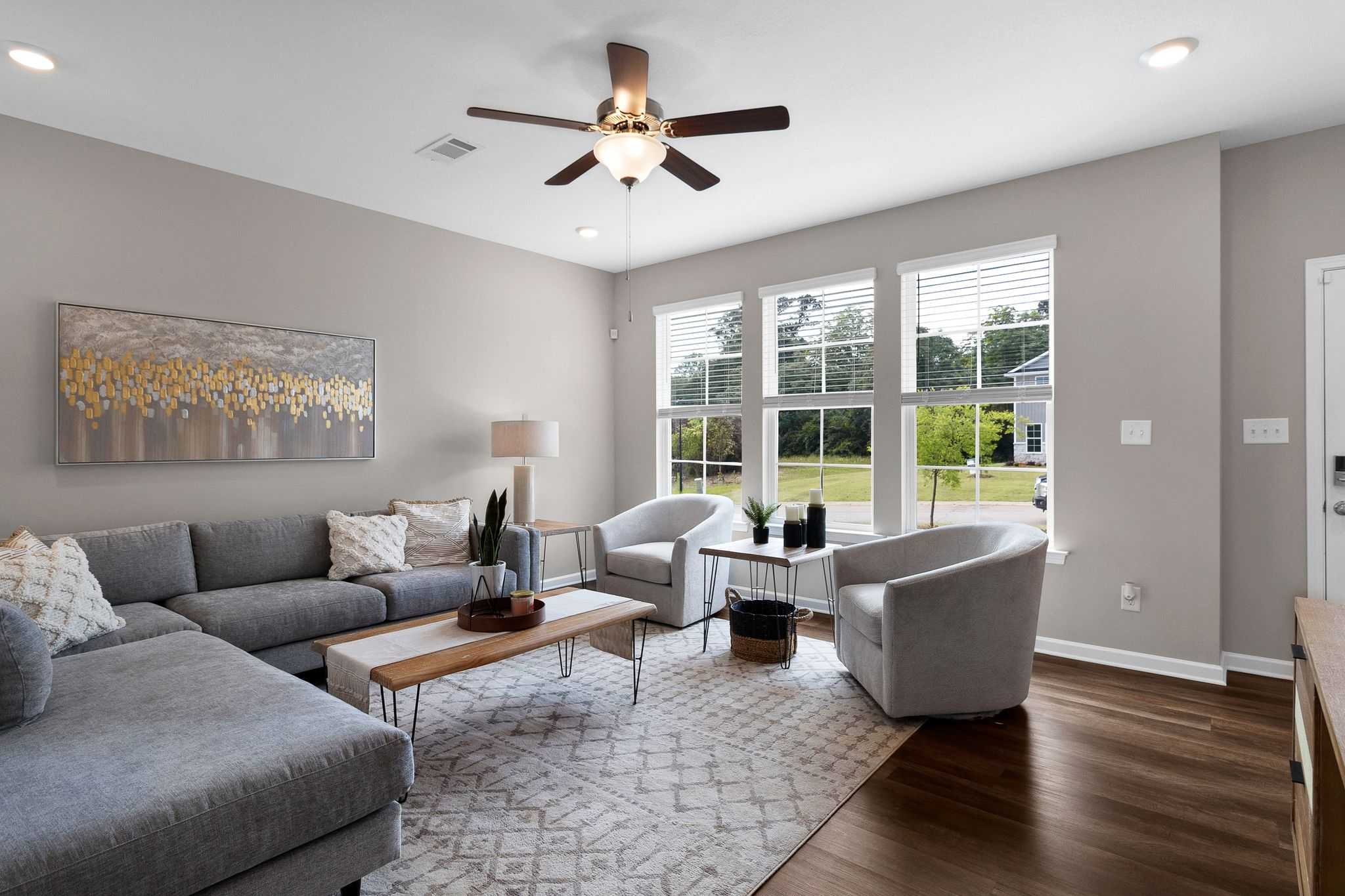 Spacious modern living room in Ivy Glen Perry GA by Evermore Homes with gray L-shaped sofa, hardwood floors, ceiling fan, large windows and abstract art