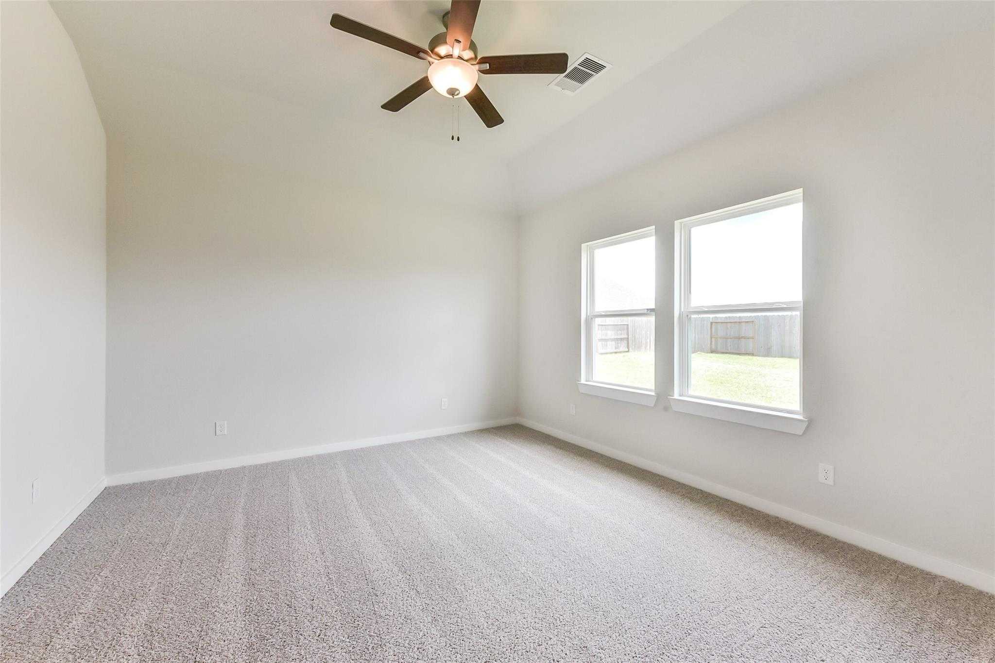 Bright bedroom with white walls, beige carpet, wooden ceiling fan, and large windows overlooking green yard in Davidson Homes The Laguna C, Rosharon, Texas