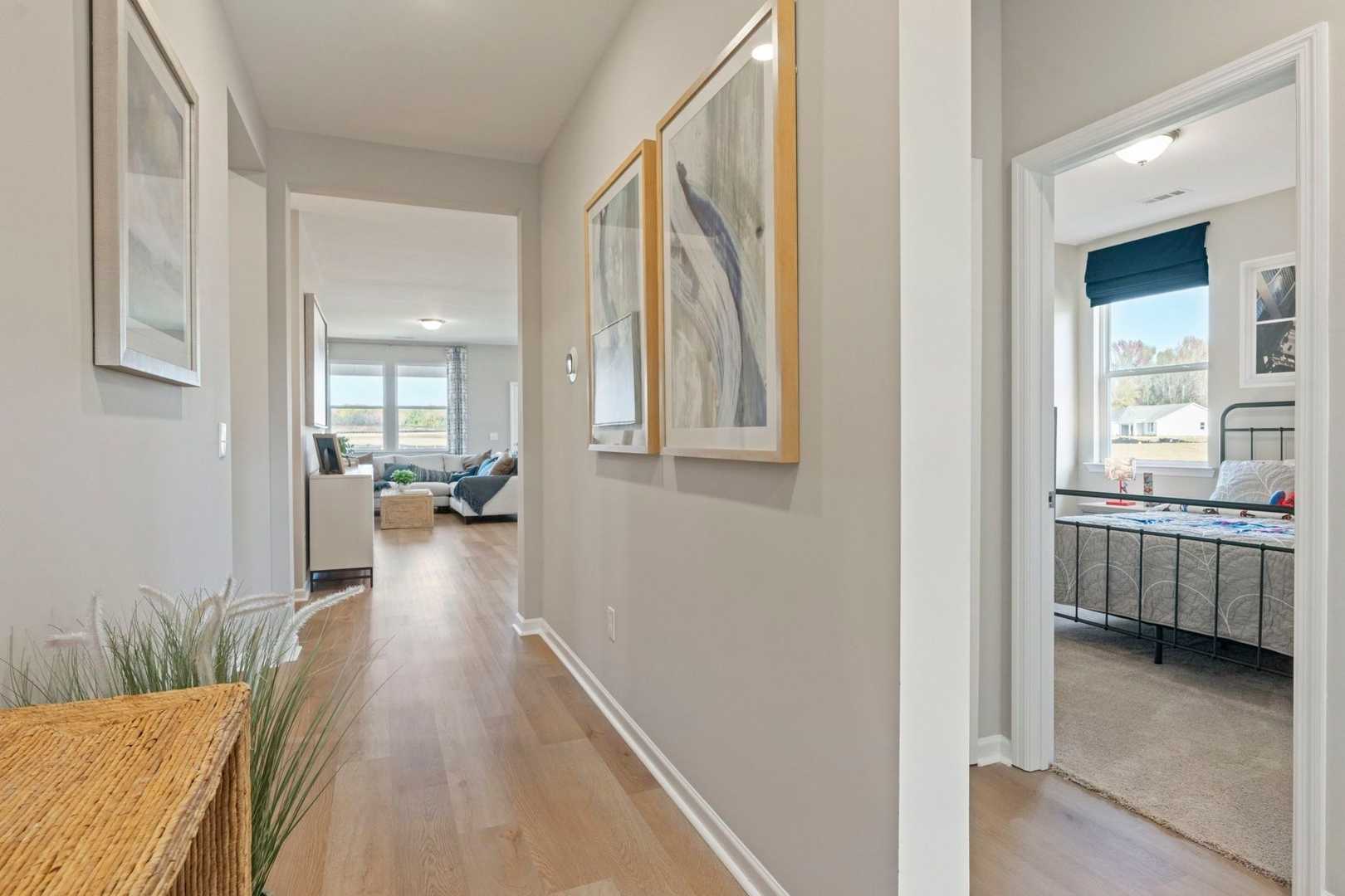 Spacious hallway in The Phoenix home design with hardwood floors, coastal art, console table, living room sofa, and adjacent bedroom with bed and blue curtains in Perry, Georgia