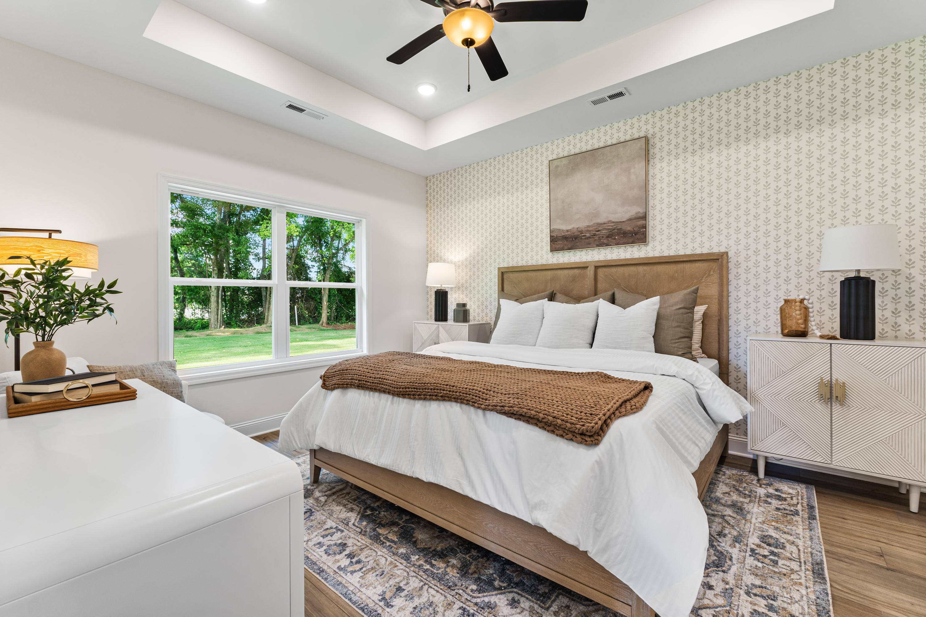 Primary bedroom at Lynn Meadows in Meridianville AL with large windows, beige upholstered bed, ceiling fan, hardwood floors