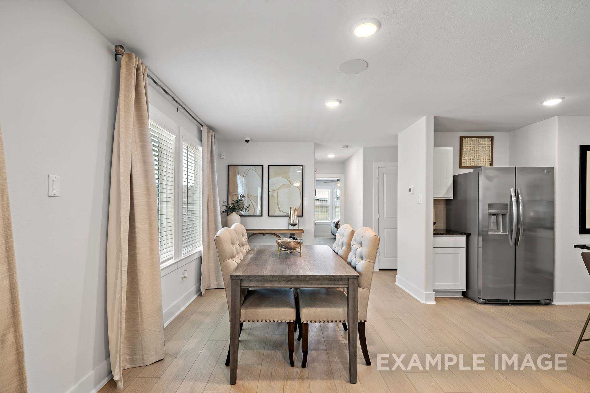 Spacious dining room in The Rose B with wooden farmhouse table, beige upholstered chairs, and open kitchen featuring stainless steel fridge