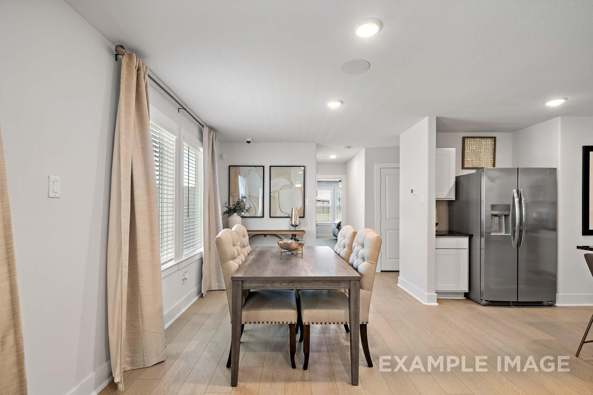 Spacious dining room in The Rose B with wooden farmhouse table, beige upholstered chairs, and open kitchen featuring stainless steel fridge